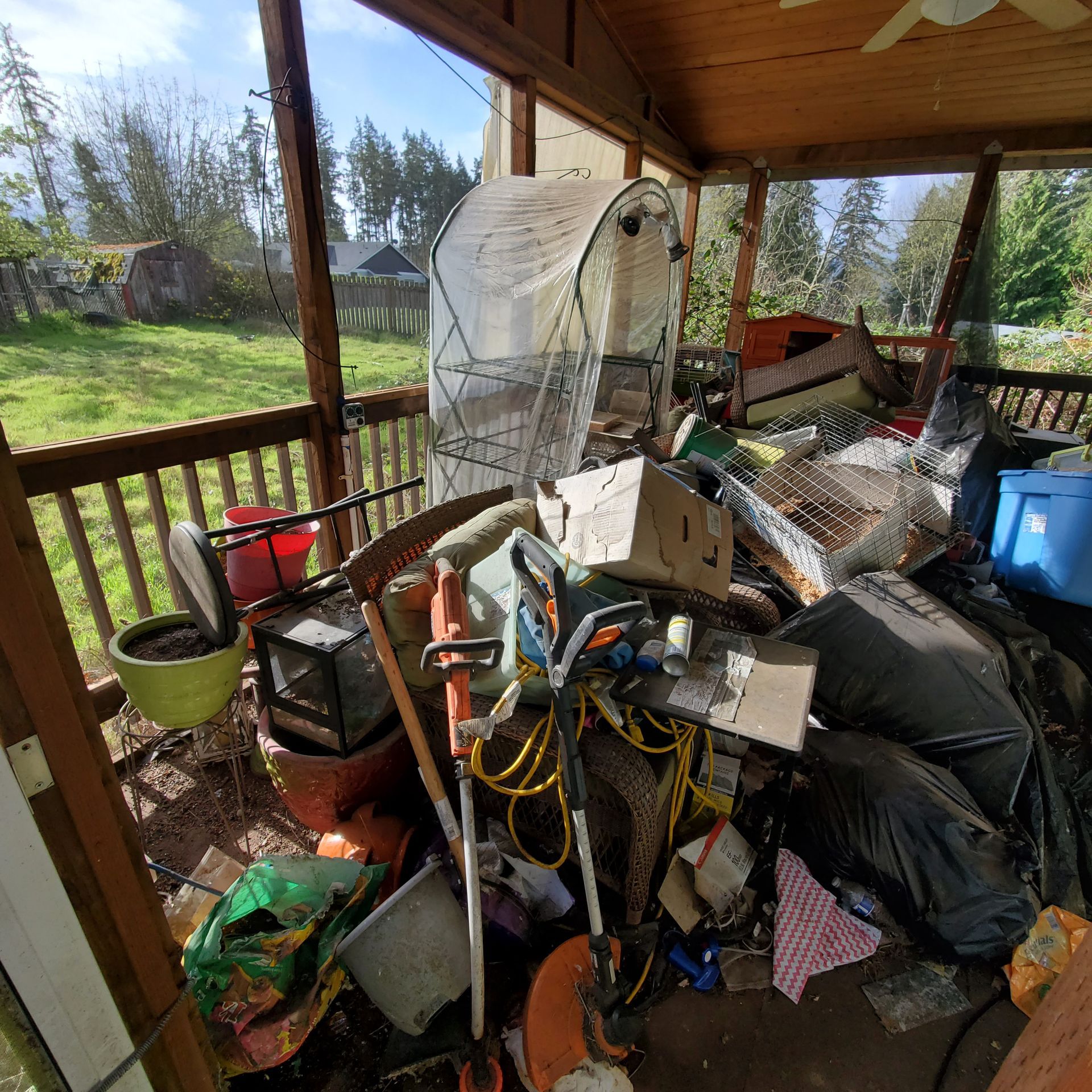 A cluttered porch with gardening tools, trash bags, and a small greenhouse.
