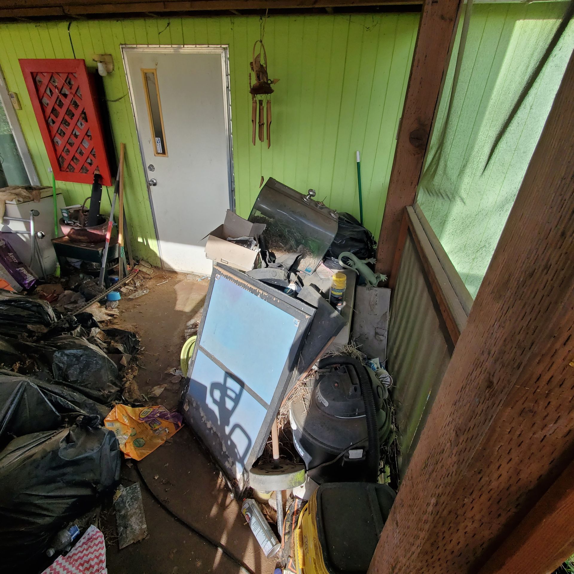 Cluttered interior with black trash bags, tools, and discarded items near a door. Green walls, daylight.
