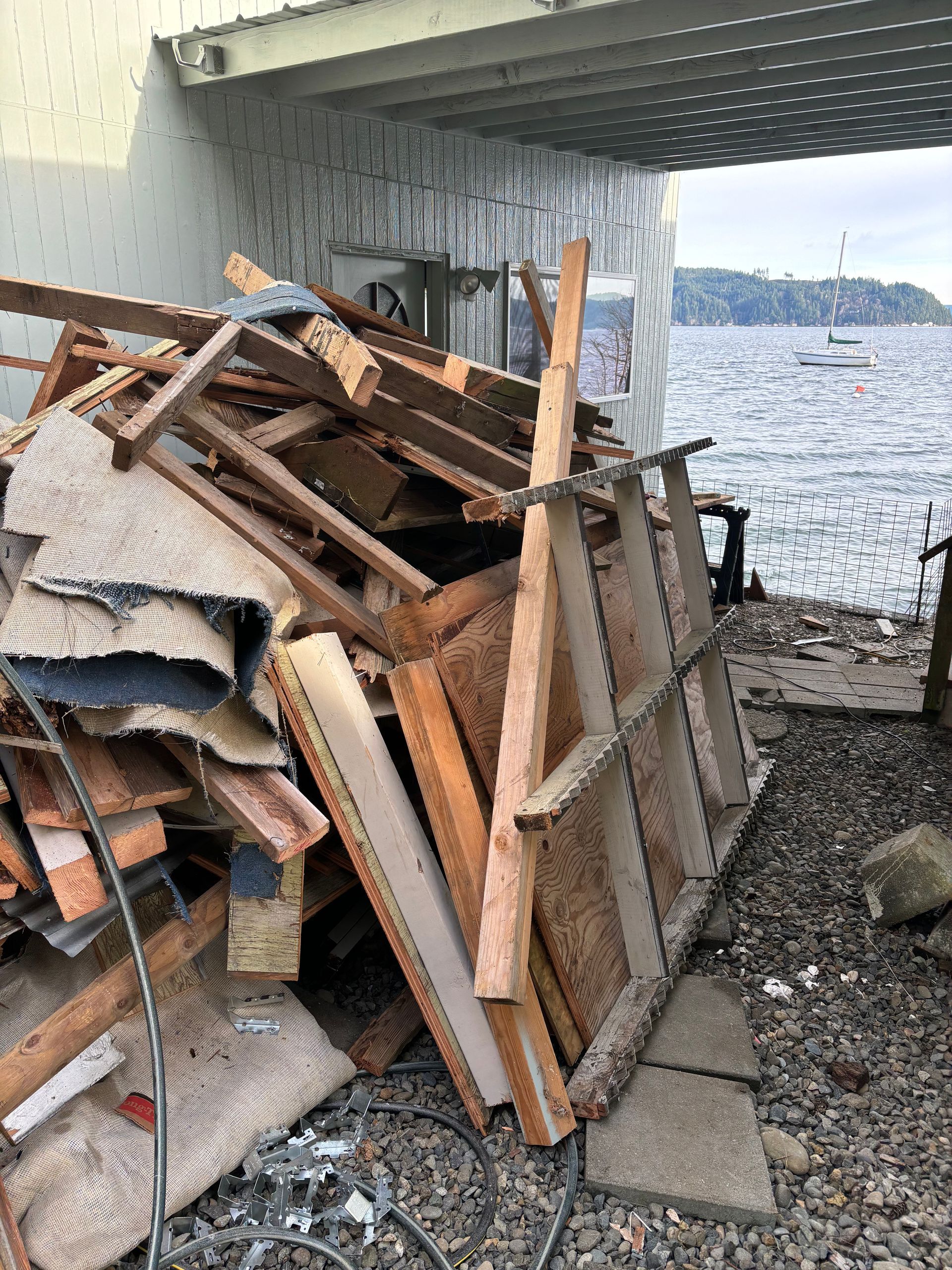 Pile of construction debris near water, beneath a bridge; includes wood, insulation, and carpet.