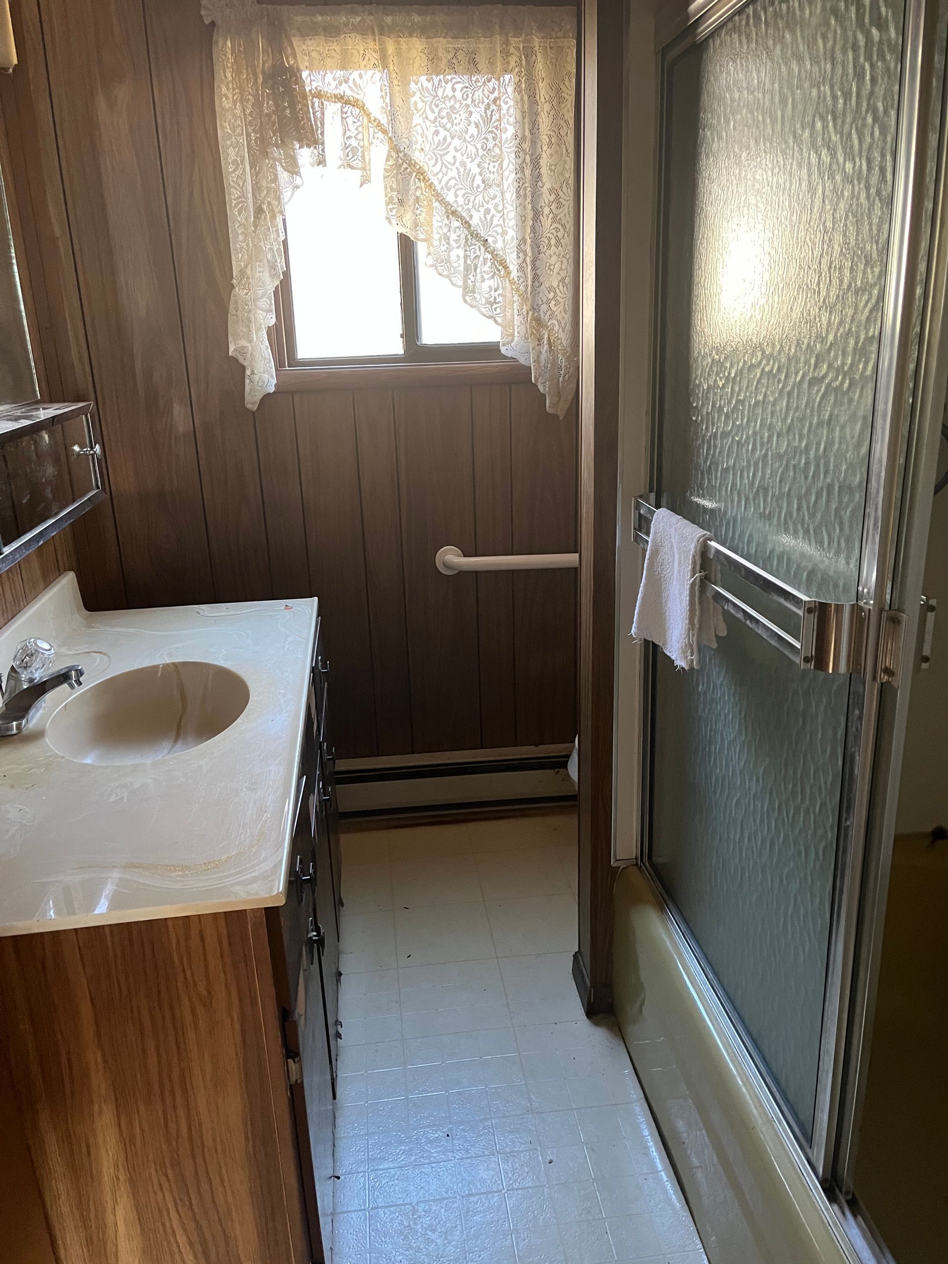 Bathroom with a vanity, a shower, and a window with lace curtains.