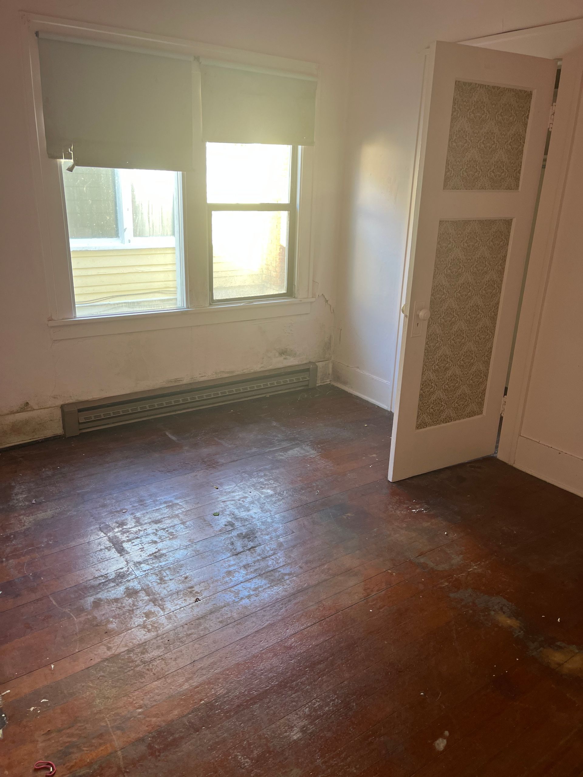 A sparsely furnished room with a window and closet, featuring stained wooden floors and peeling paint.