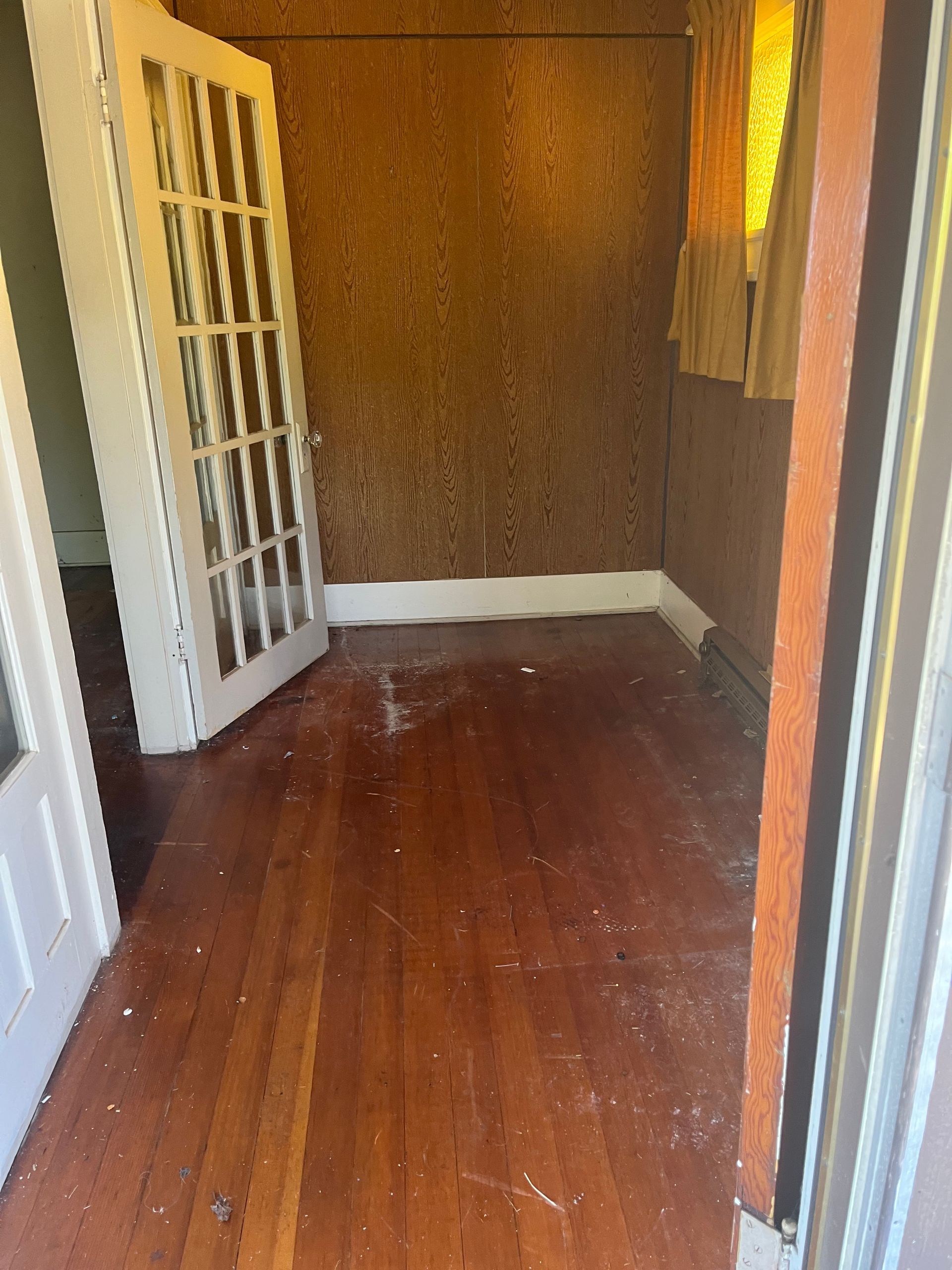 Room with wood floor, brown walls, white French door, and yellow-curtained window.