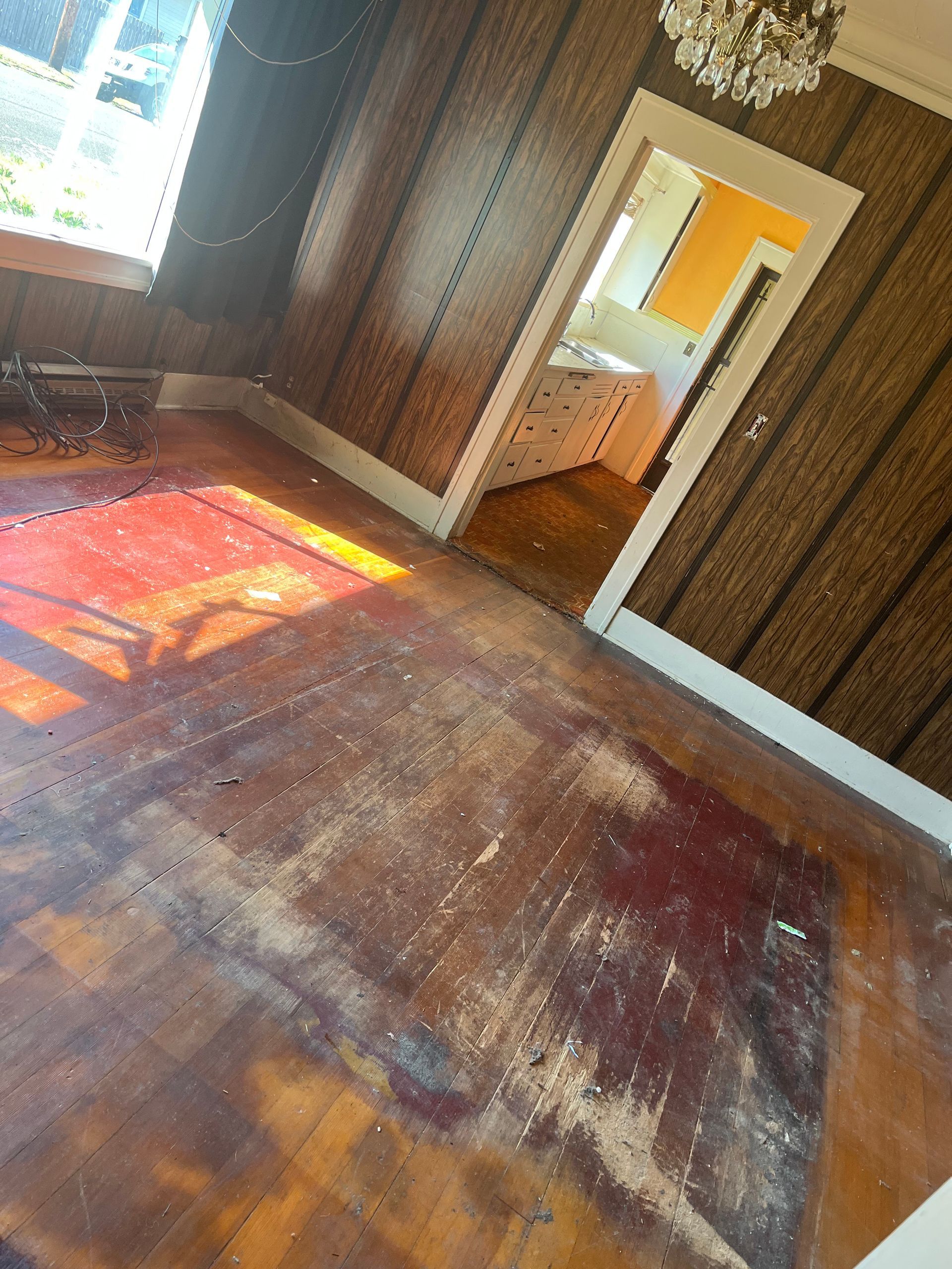 Wooden floor with stains in a room with textured brown wallpaper and a doorway leading to a white cabinet.