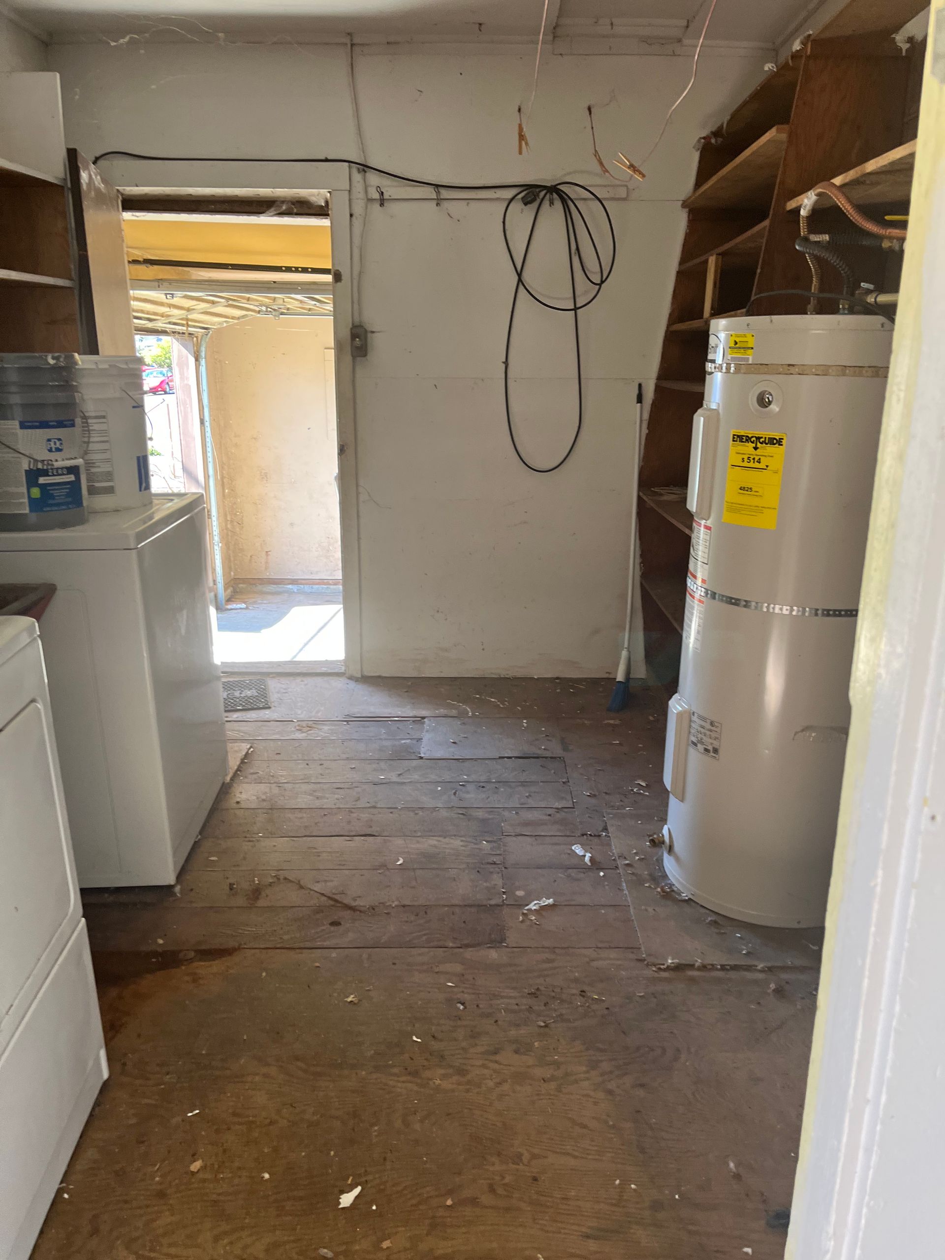 Laundry room with water heater, washer, and doorway to outside. Dirty floor, white walls.