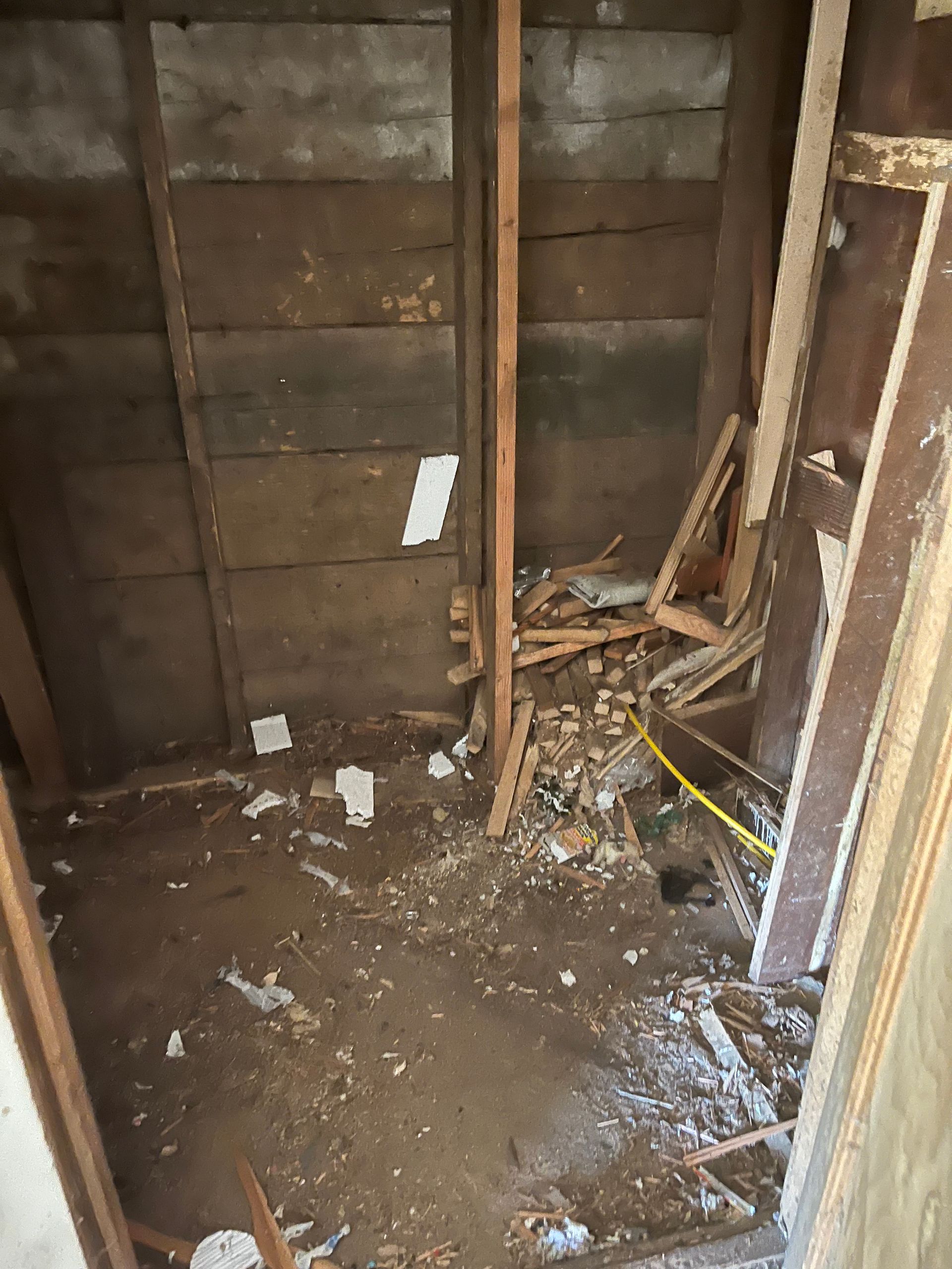 Interior of a room under renovation, revealing wooden frame and damaged walls. Debris covers the floor.