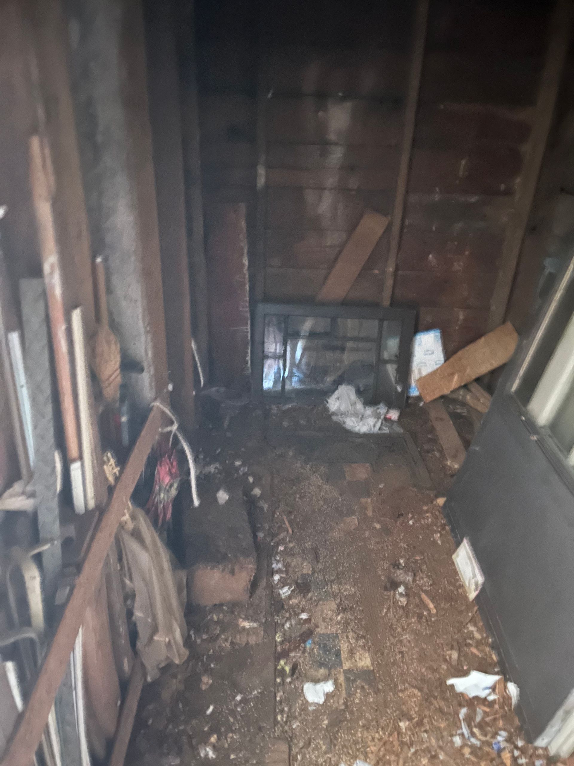 Dirty, cluttered storage space; wood walls and floor; debris and materials piled up.
