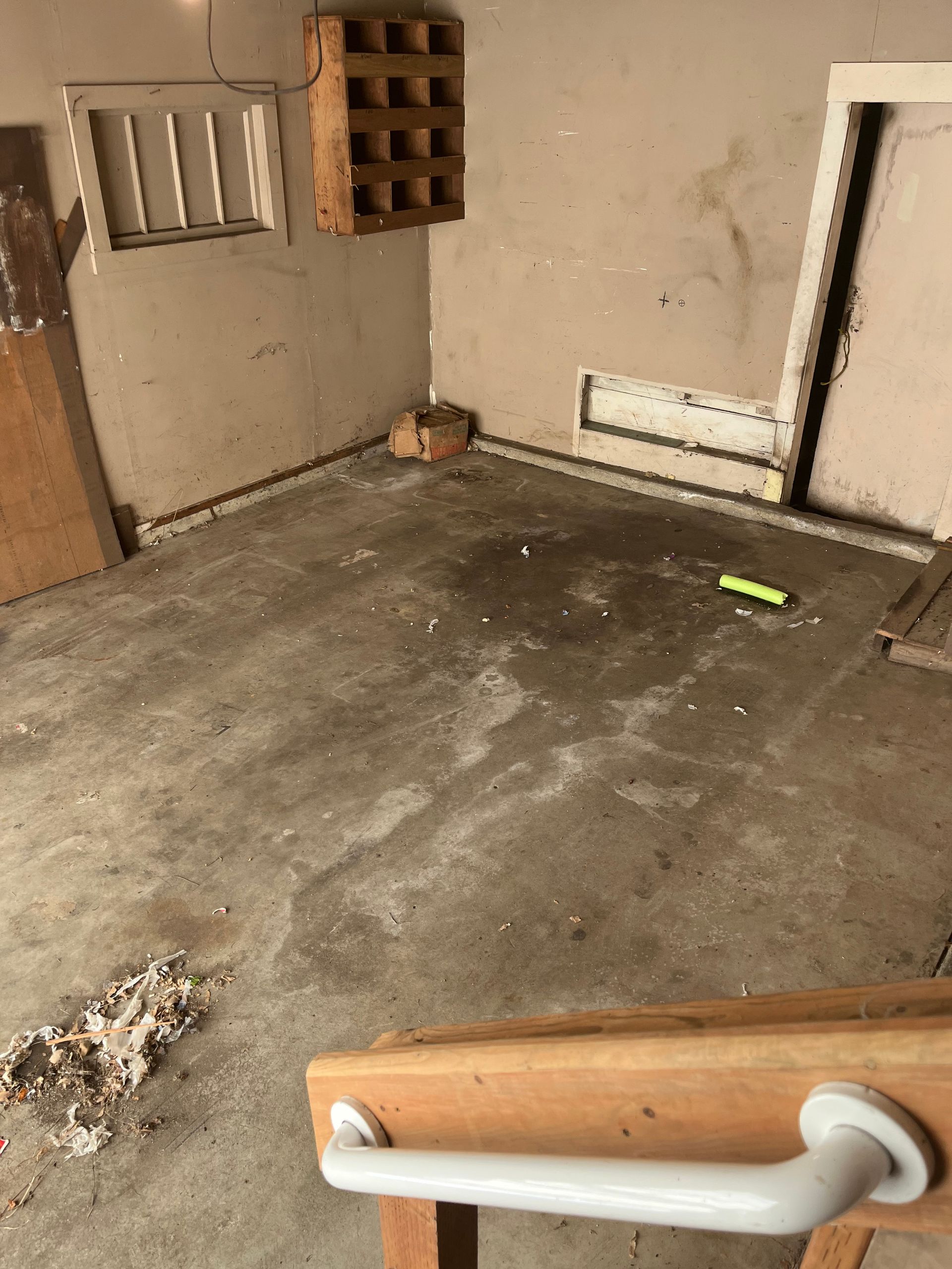 A cluttered, dingy room with concrete floor.  A wooden wine rack hangs on the wall, a white door.