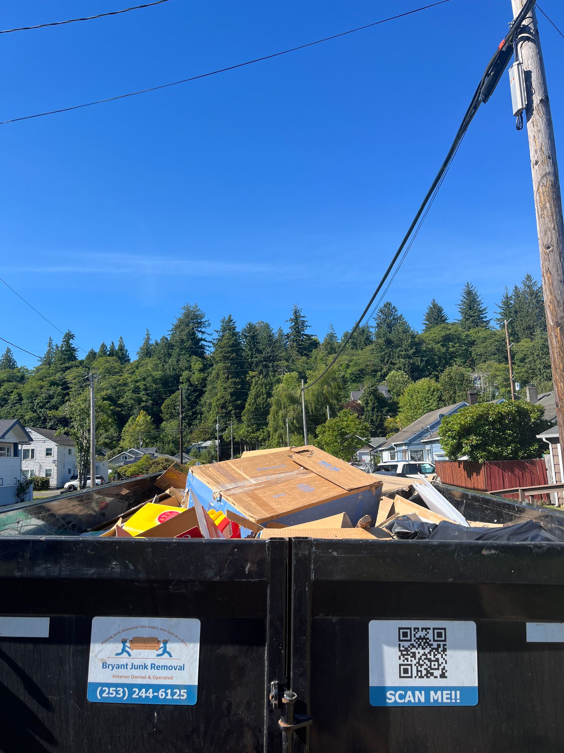 Dumpster overflowing with construction debris against a backdrop of houses and a forest. Blue sky overhead.
