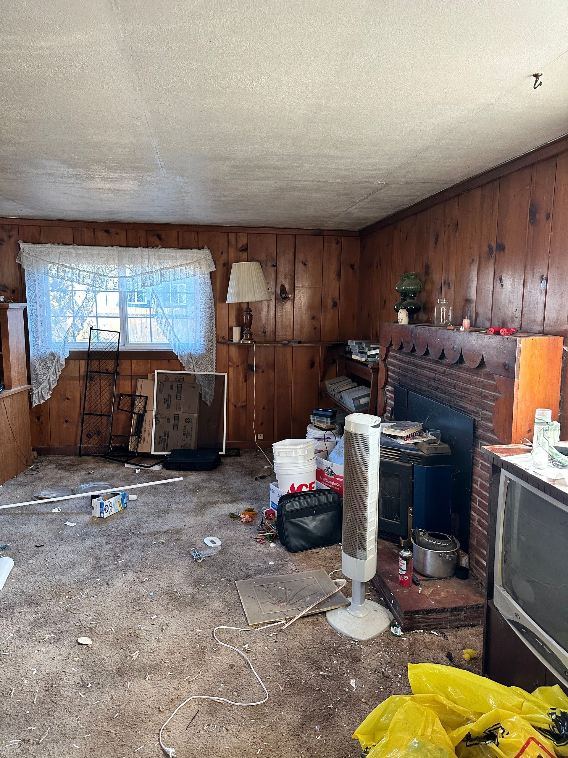Messy living room with wood paneling, cluttered floor, and a damaged fireplace.