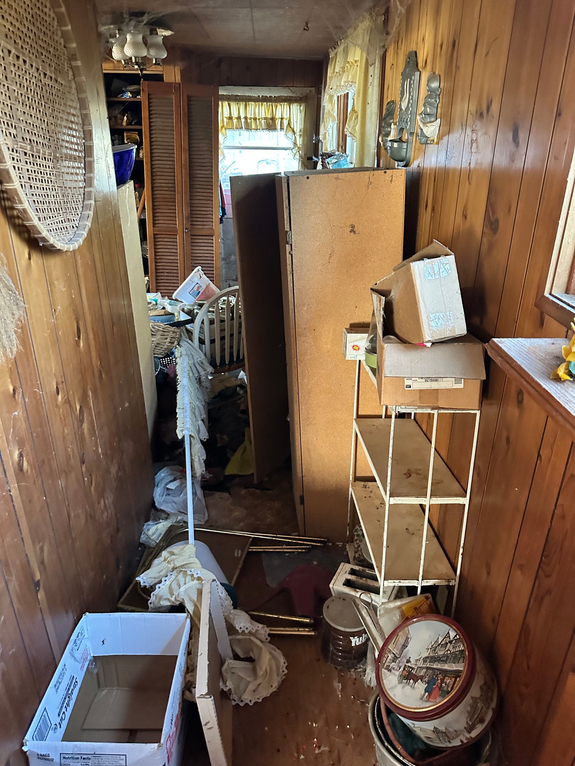 Cluttered hallway with wood paneling, boxes, shelves, and debris, obstructing passage.