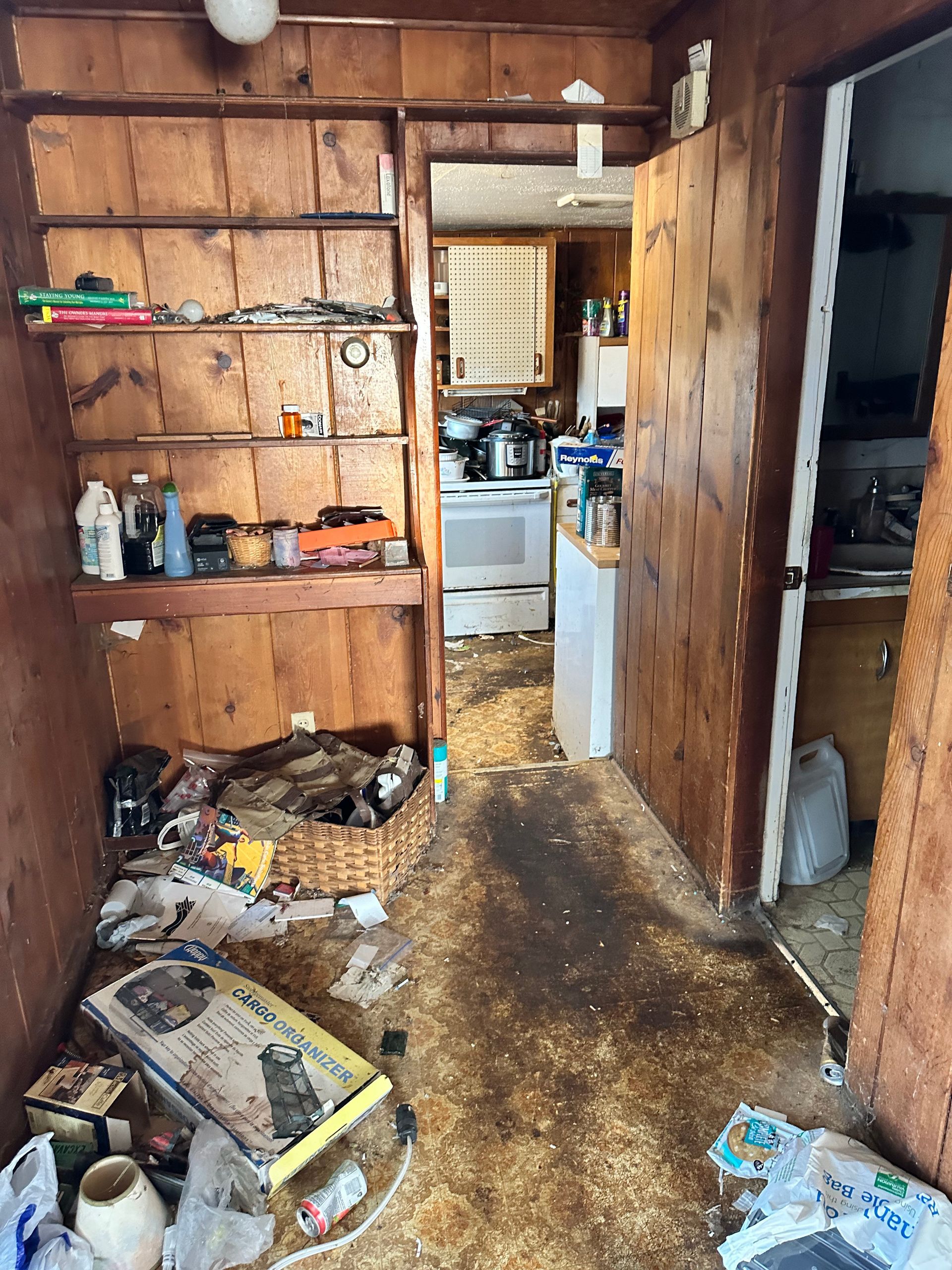 Cluttered room with wood paneling, shelves, and doorway to a kitchen. Debris litters the floor.