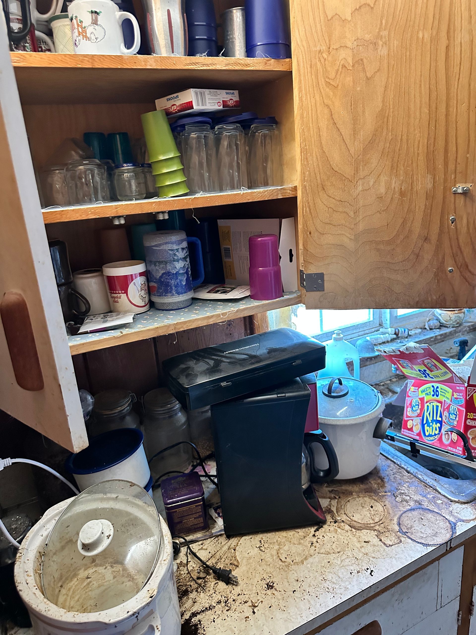 Kitchen cabinet with dishes, and countertop with appliances and debris.
