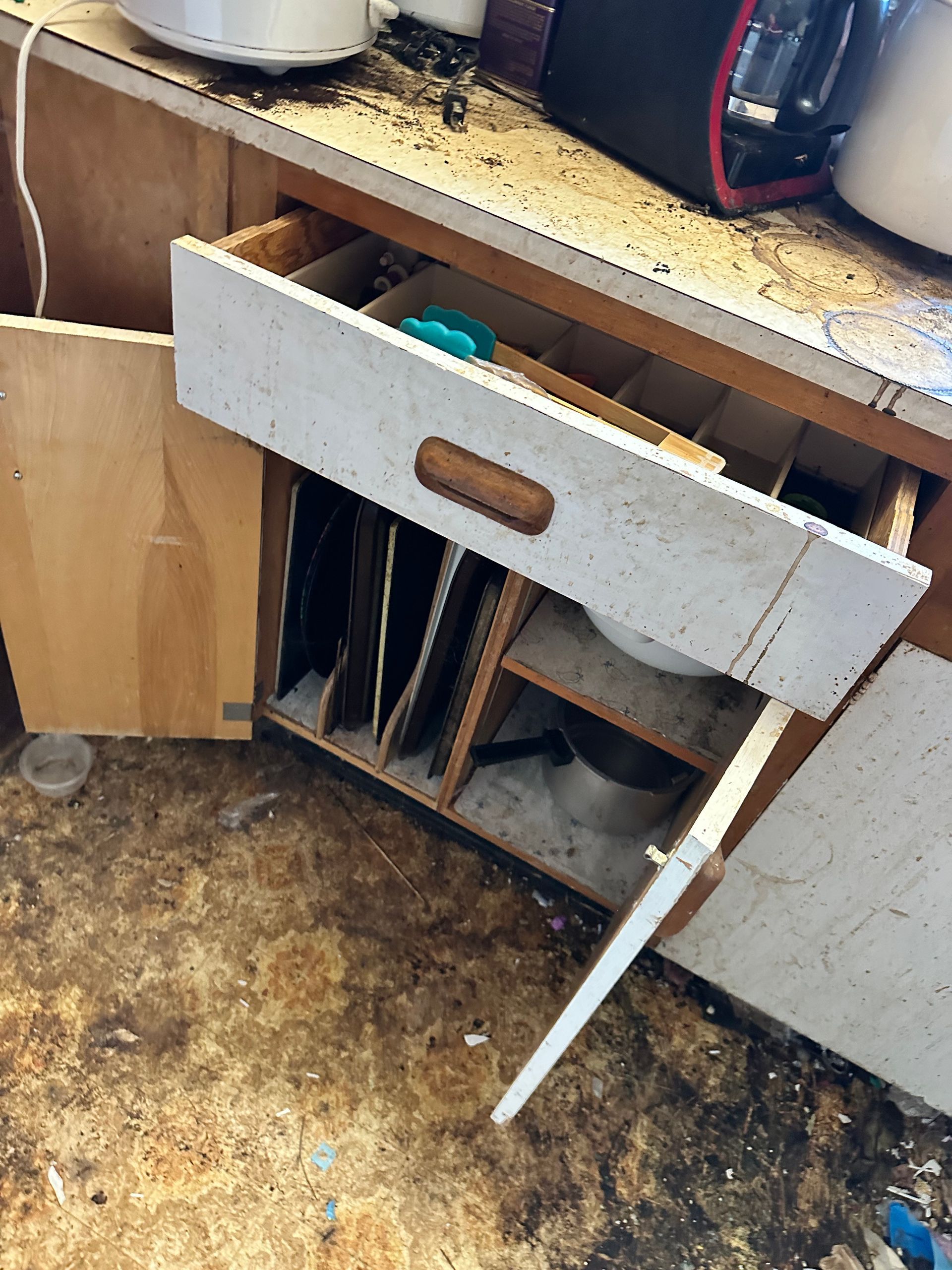 Open kitchen cabinet with a drawer pulled out, dirty floor and contents.