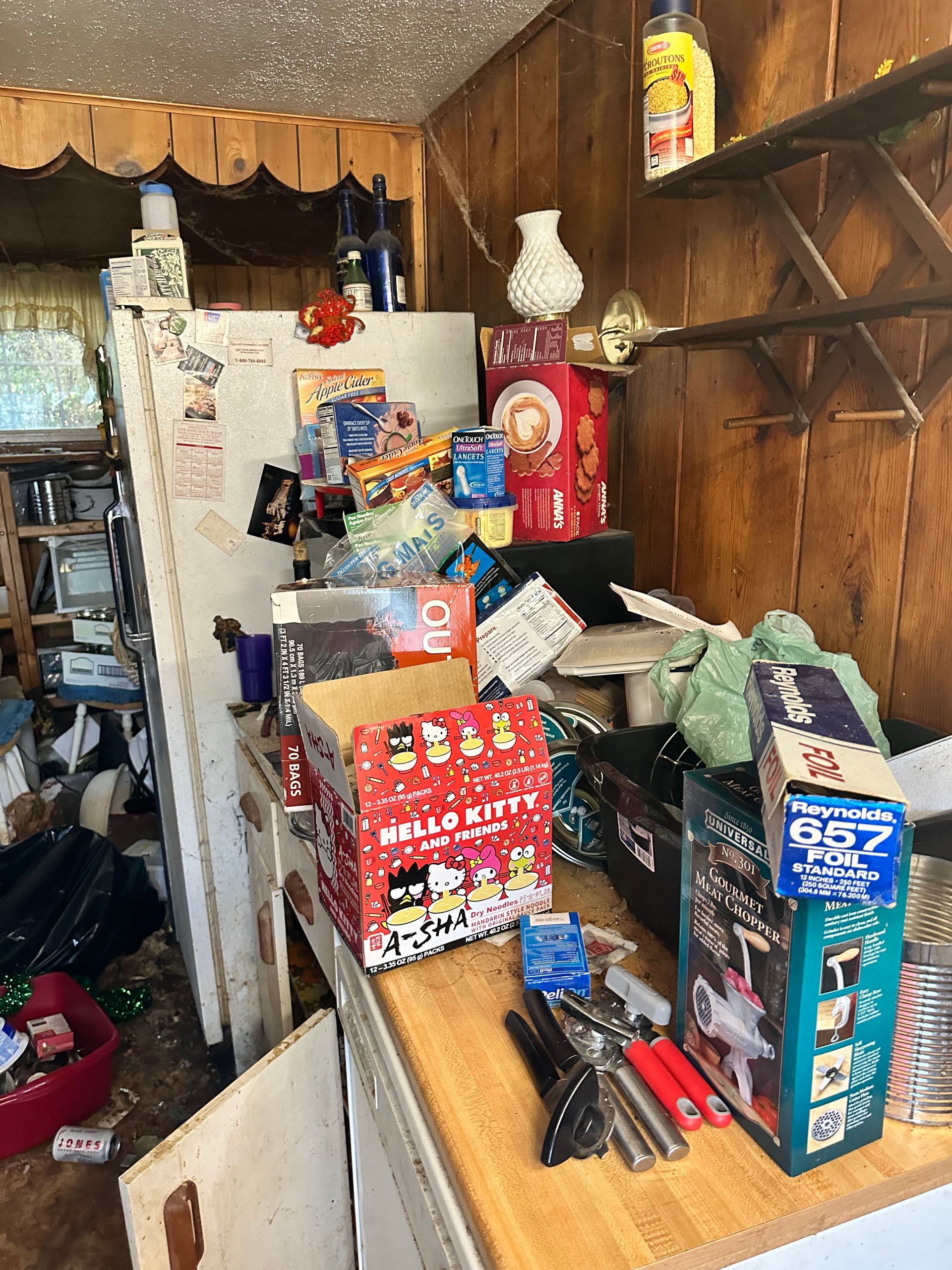Cluttered kitchen: Fridge covered in items, boxes, and a collection of objects on the countertop, with wooden paneling.