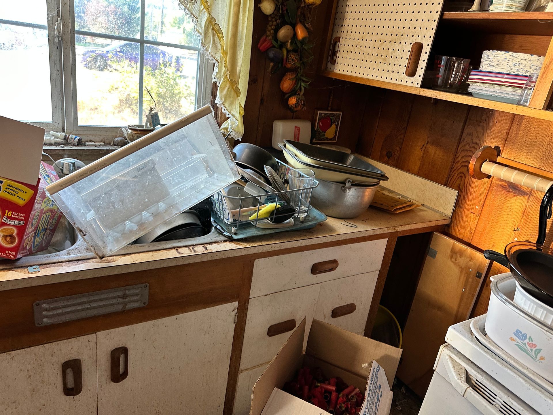 Cluttered kitchen counter with dishes, boxes, and appliances, beside a window with an outside view.