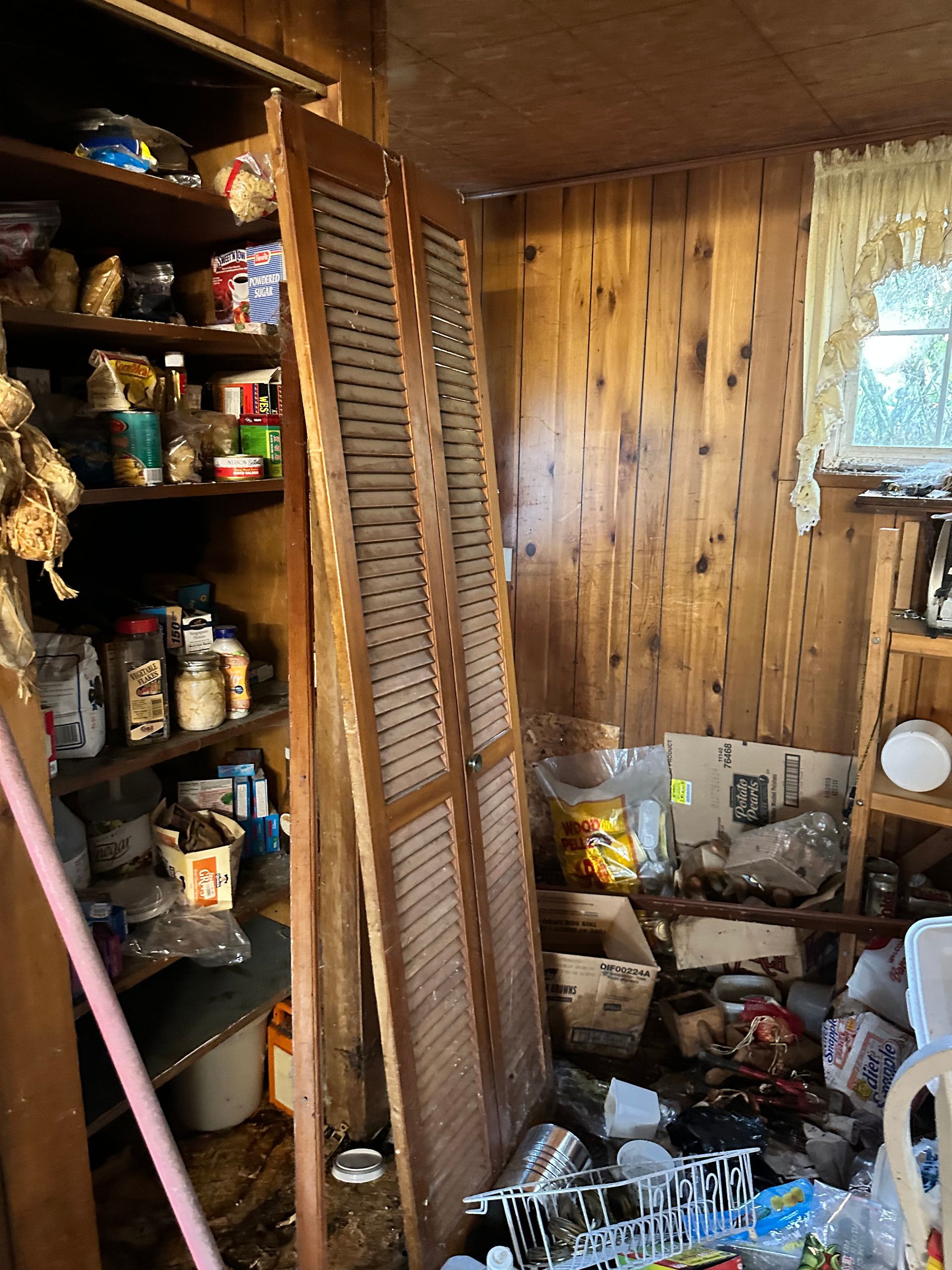 Cluttered pantry with wooden shelves and doors. Shelves hold various food items and debris.