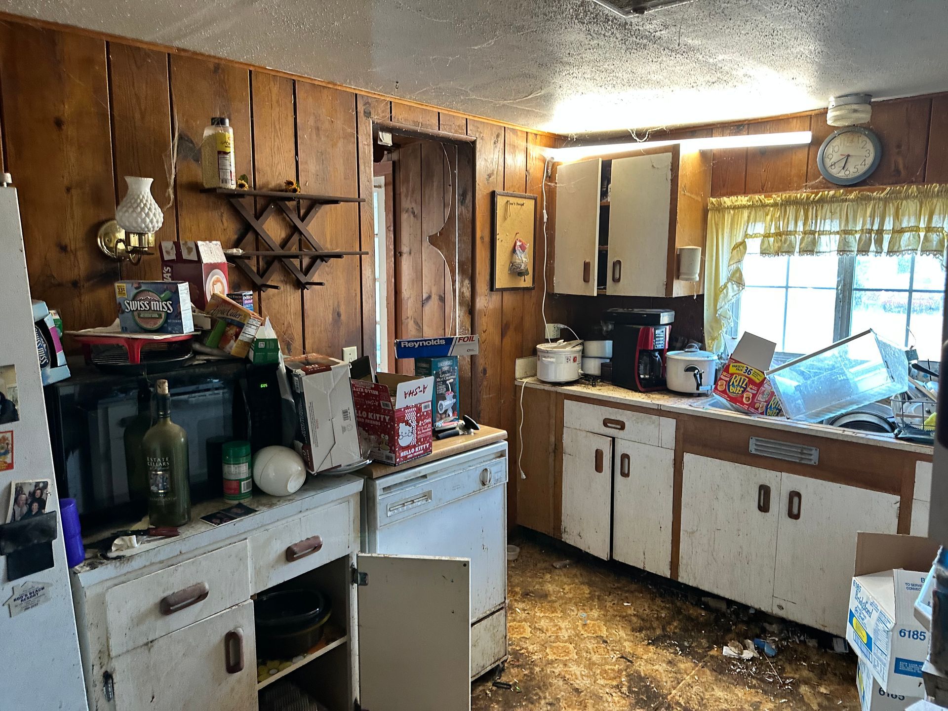 Cluttered kitchen with old wooden cabinets, appliances, and various items; damaged walls.