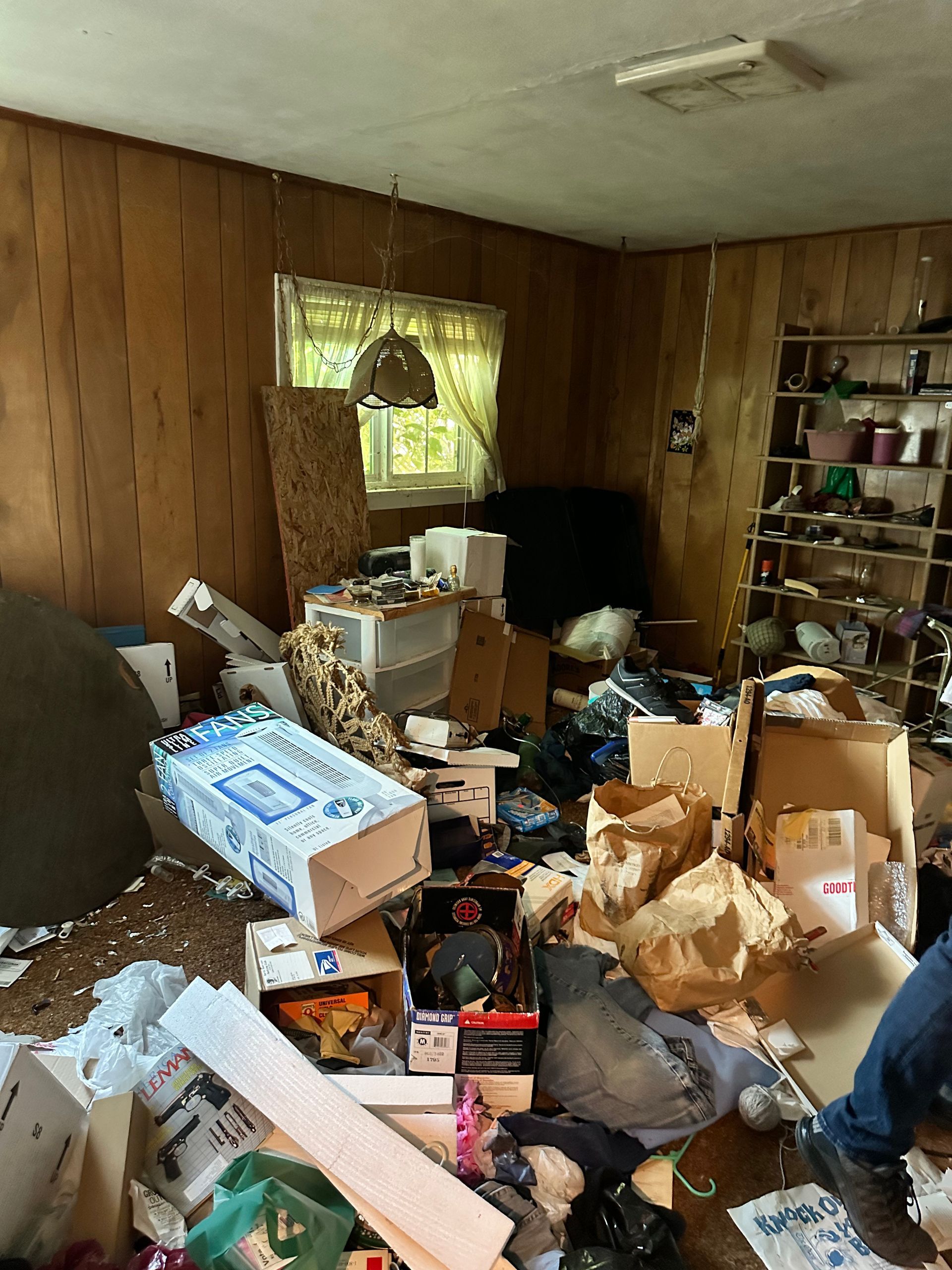 Cluttered room with boxes and debris. Wood-paneled walls, window with sheer curtain, shelves on right side.