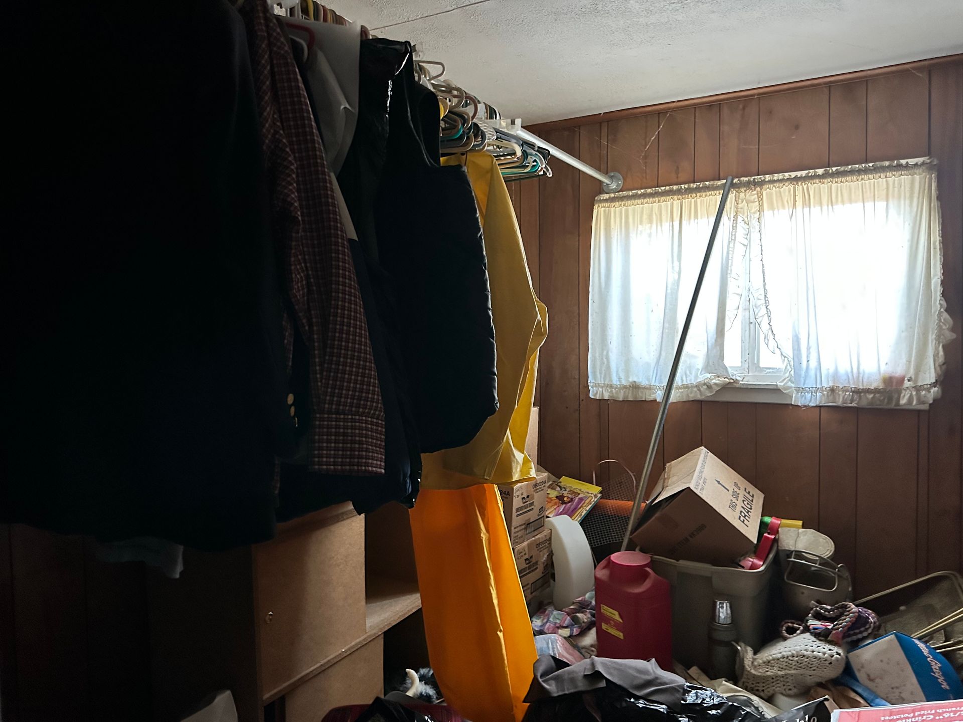 Clothes hanging in cluttered room with a window, boxes, and debris on the floor.