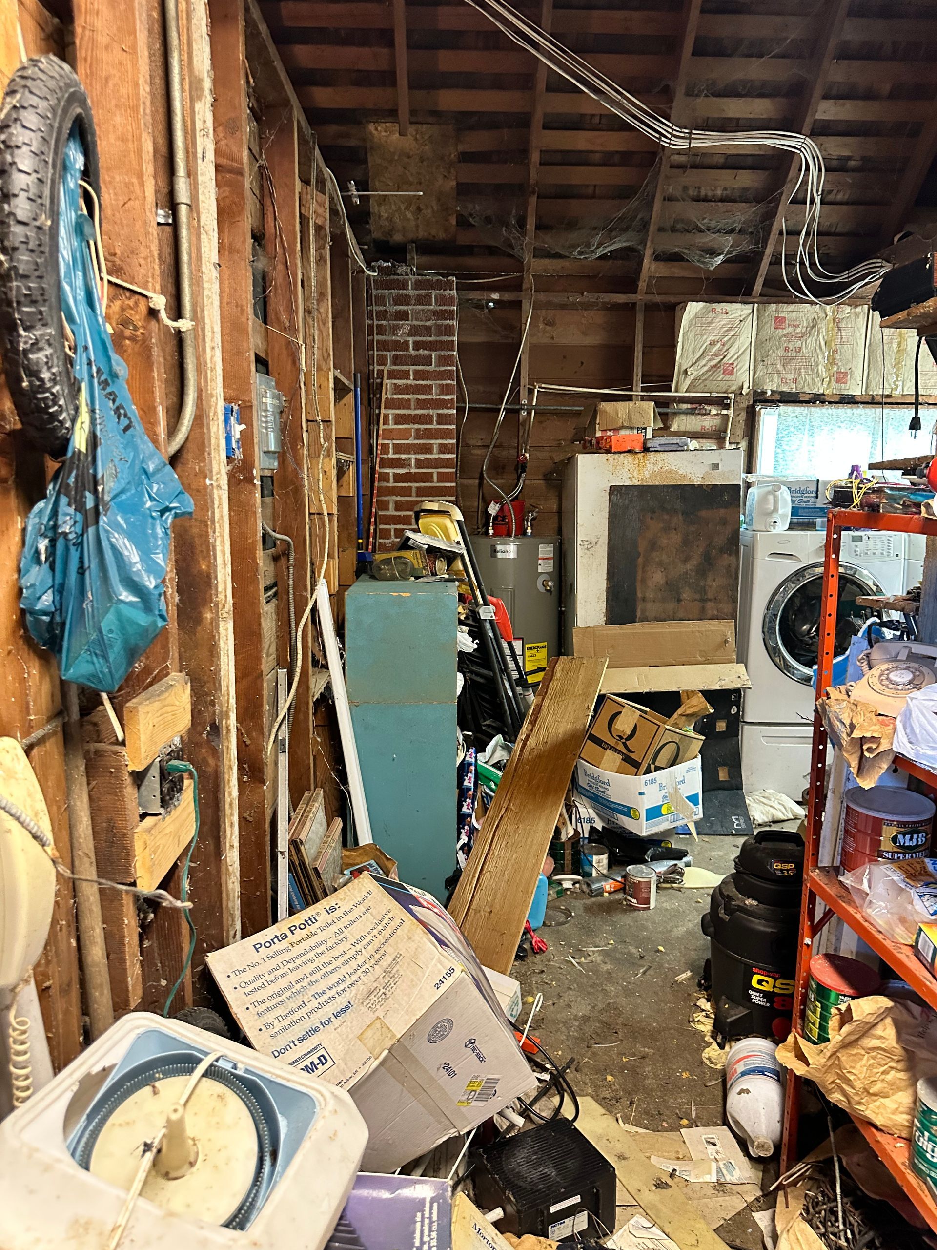 Cluttered, messy garage interior with tools, debris, appliances, and exposed wood beams.