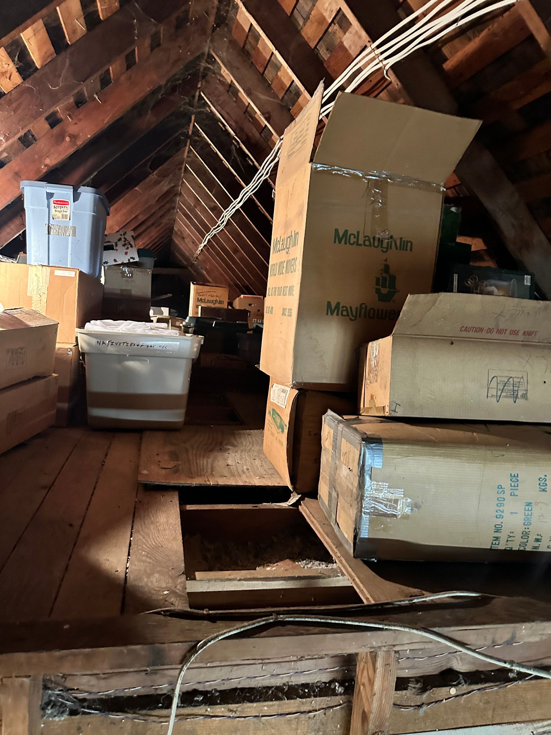 Cluttered attic with boxes and exposed beams; a hole is in the wooden floor.