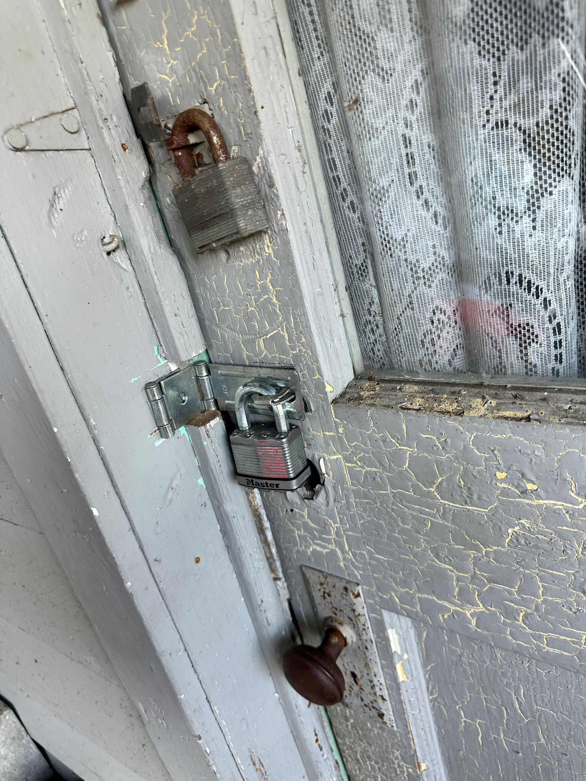 A locked, old, white door with a screen, featuring a knob and two padlocks.