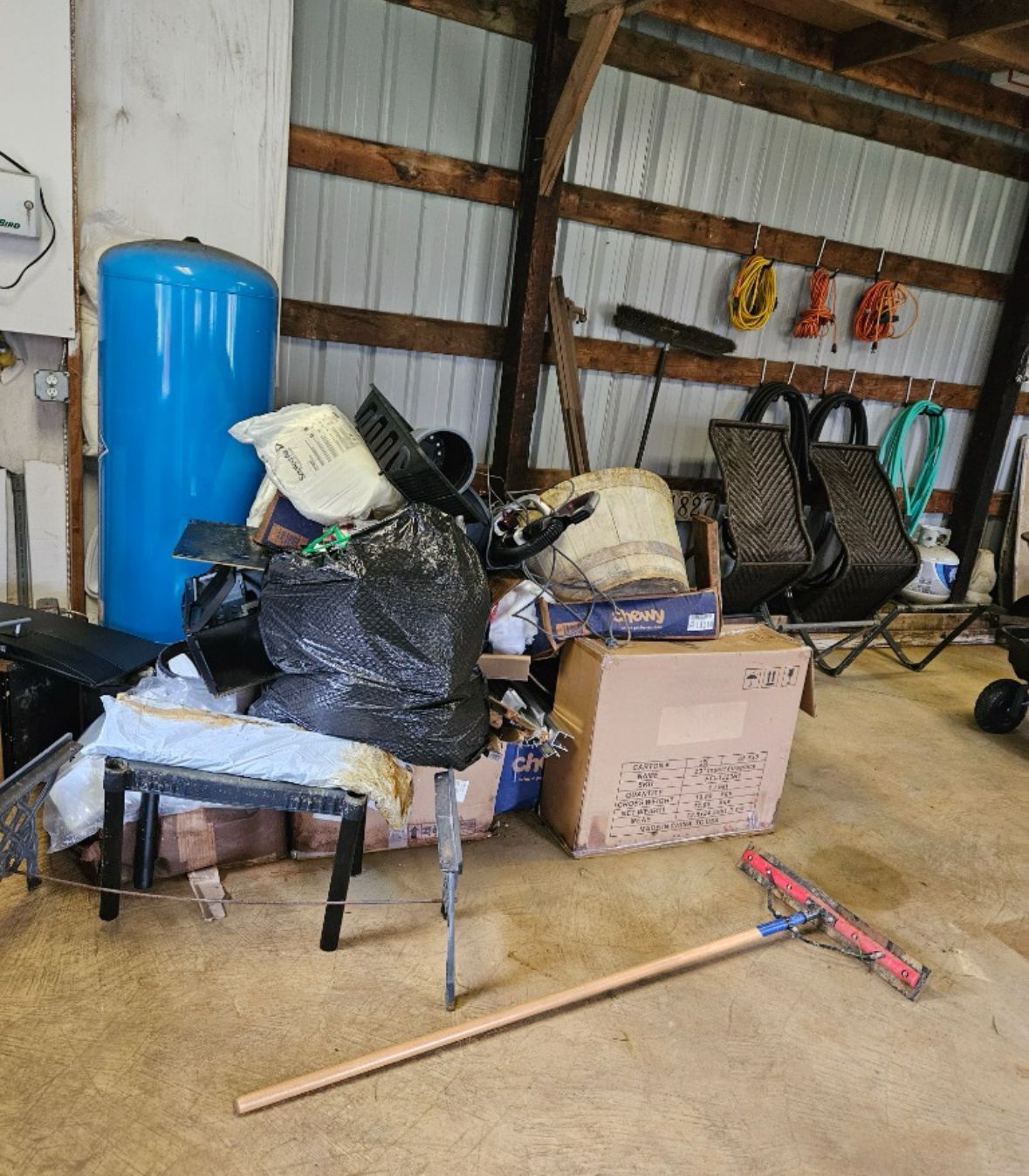 Cluttered shed interior: blue water tank, trash bags, boxes, lawn rake, and assorted items.
