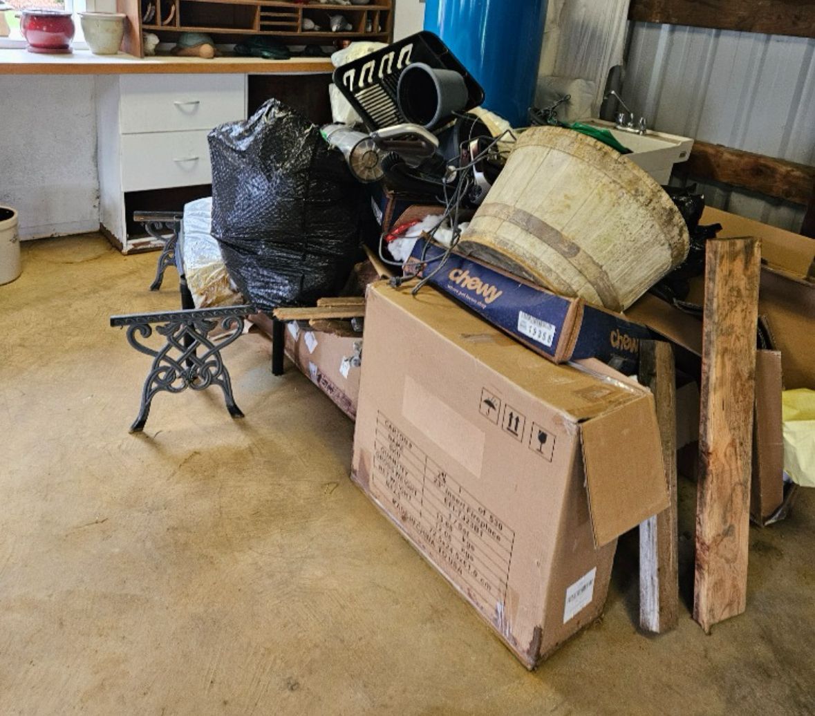 Jumbled pile of trash and discarded items in a cluttered room. Includes boxes, wood planks, and a decorative metal table leg.