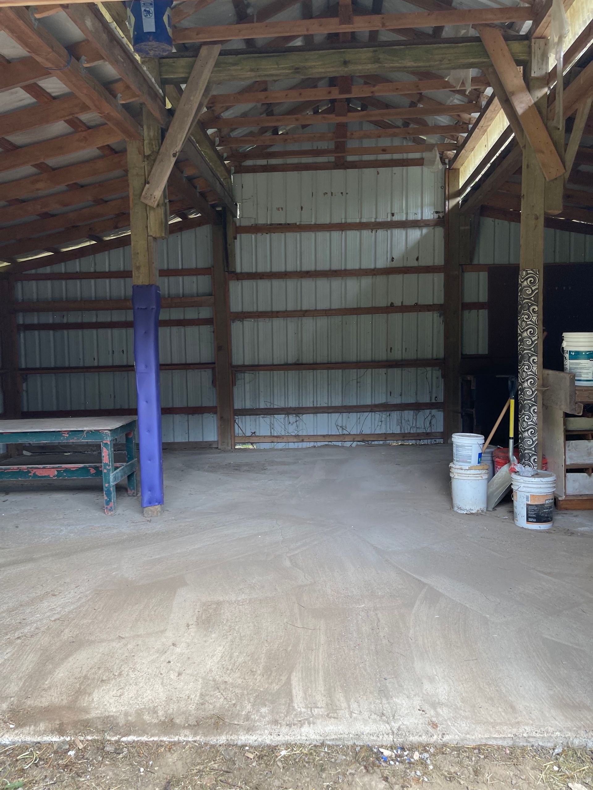Interior of a barn. Gray concrete floor, metal walls, wooden beams. Buckets of paint on the right.