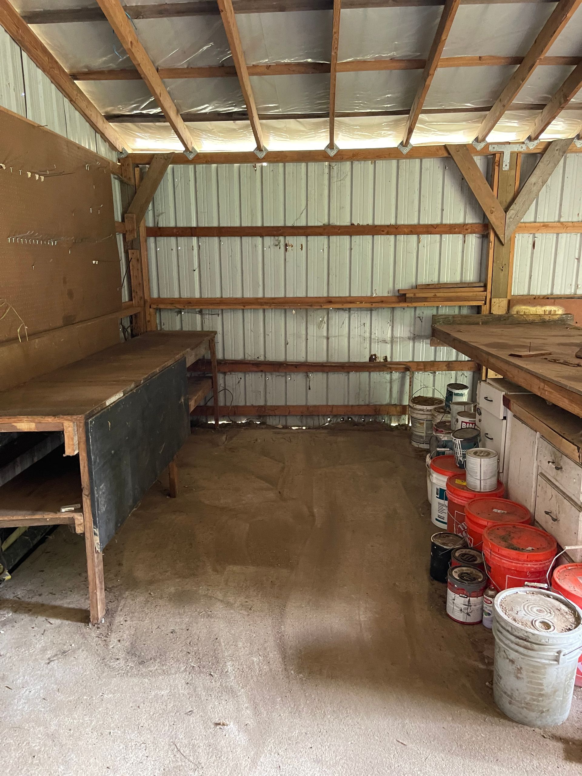 Interior of a workshop with two workbenches, storage, and paint cans. Wooden structure, metal siding.