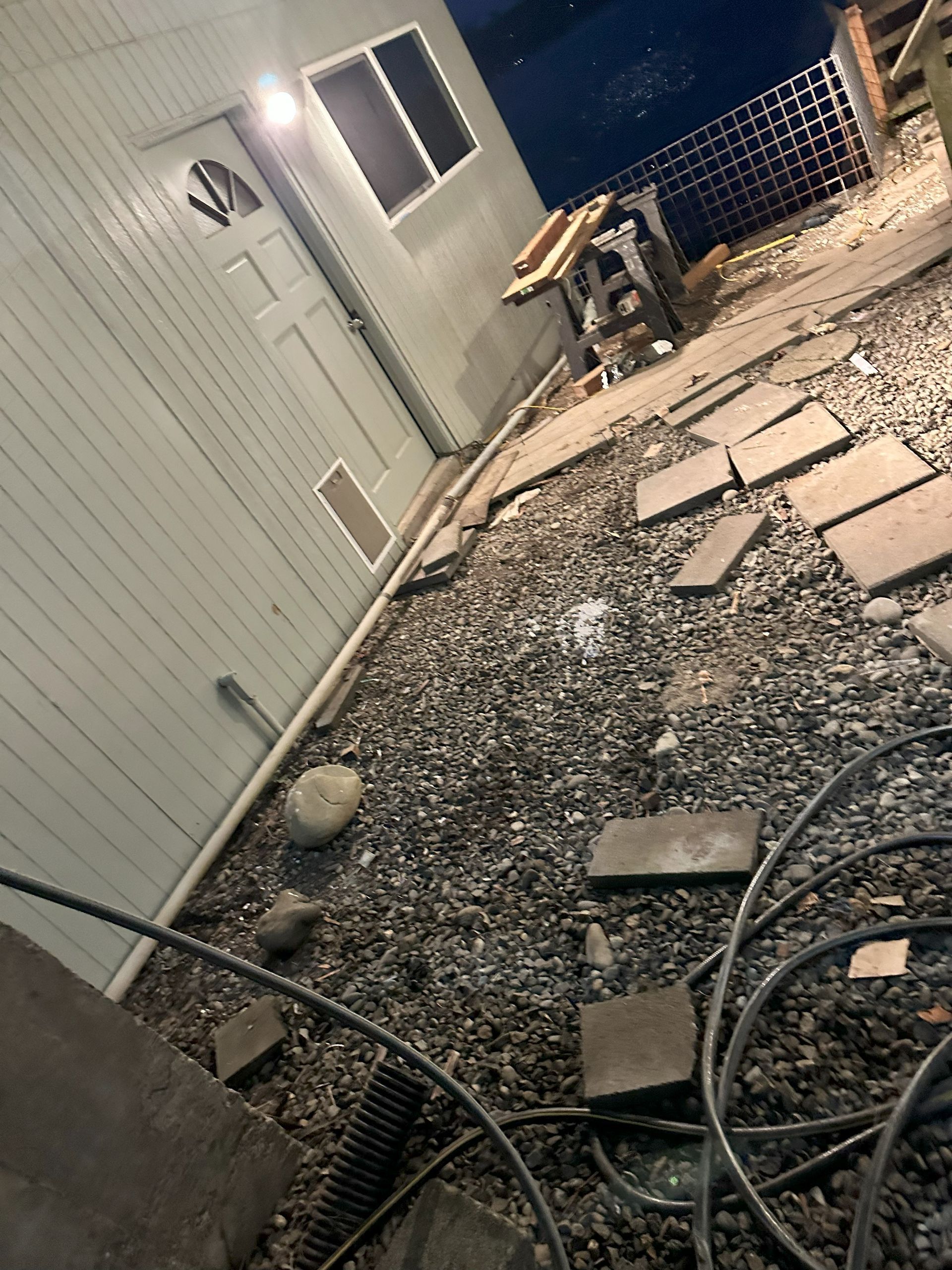 Exterior: Gravel yard with concrete stepping stones, a building with a door and window, and electrical cords.