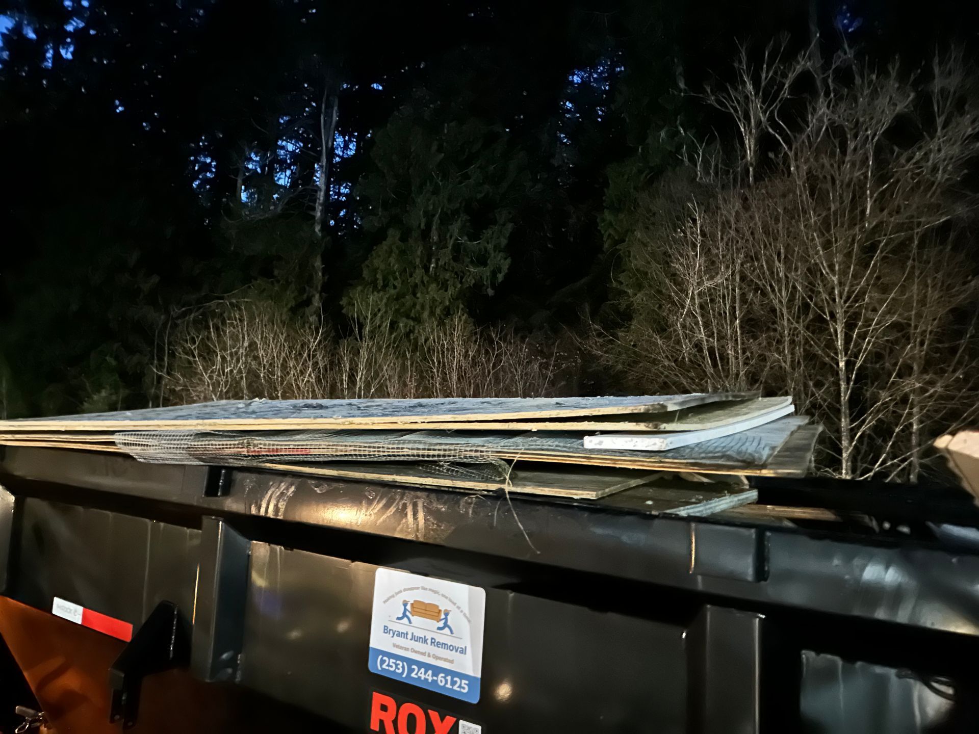 Dumpster loaded with wood debris at night, with a forest background.