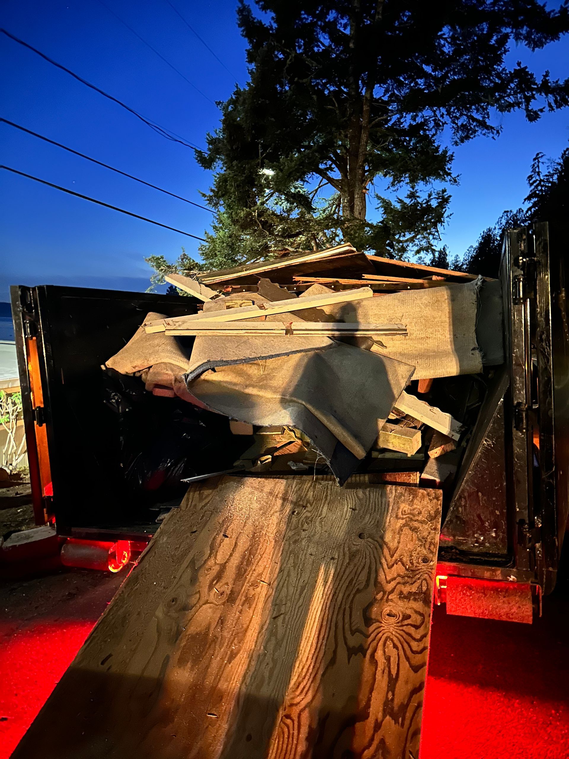 A debris-filled trailer with a wooden ramp, illuminated by red light, against a twilight sky.