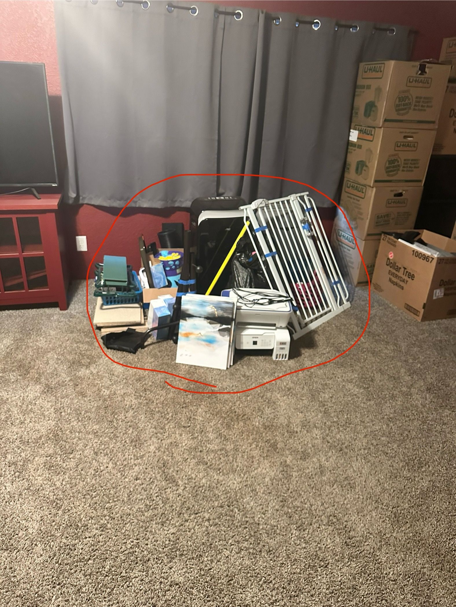 Cluttered pile of equipment on carpet, including a printer, mixing board, and other electronics, near a red wall and curtains.