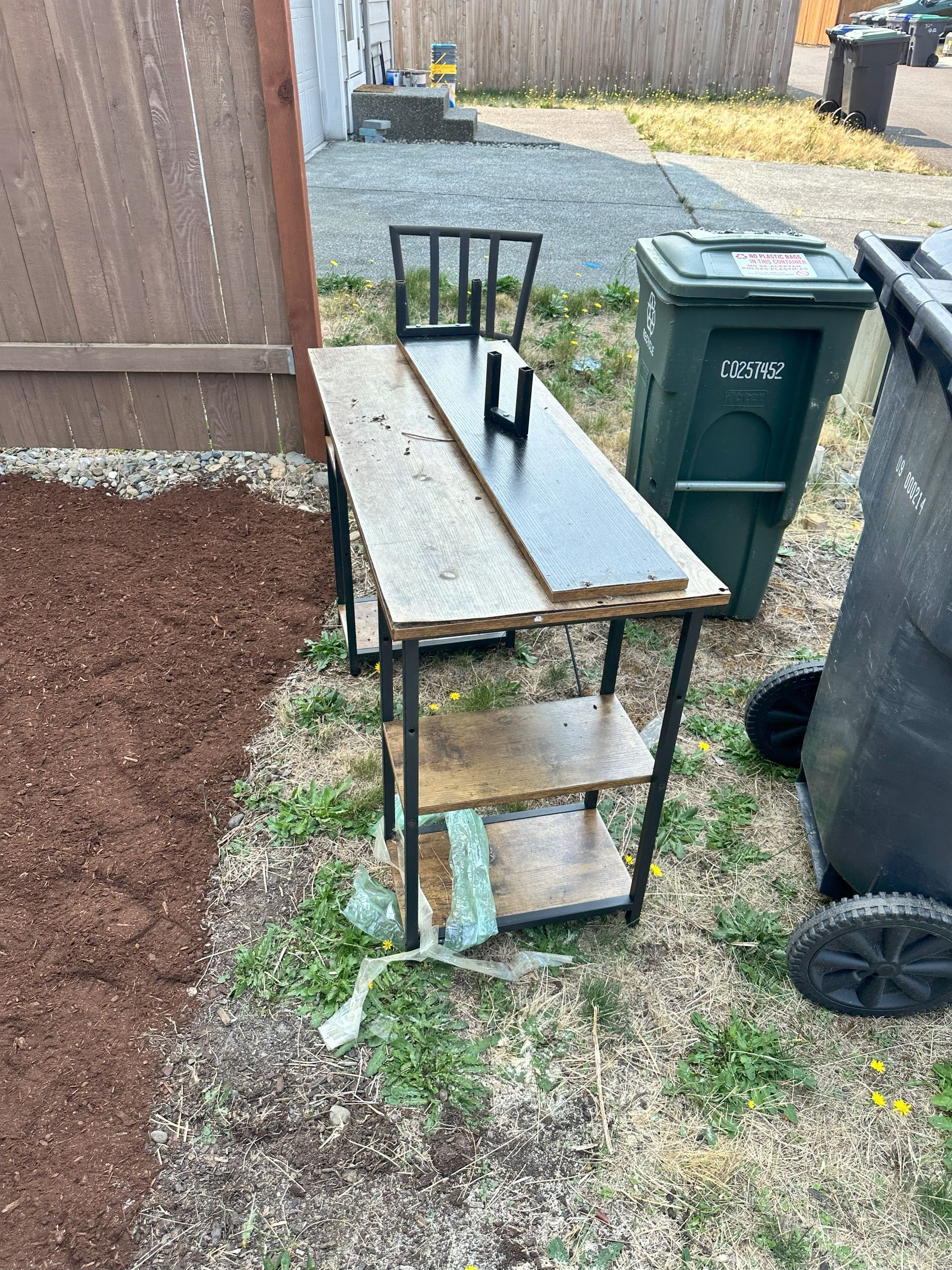 Table with a chair and garbage cans. The table has shelves with various wood planks. Outdoors on grass.