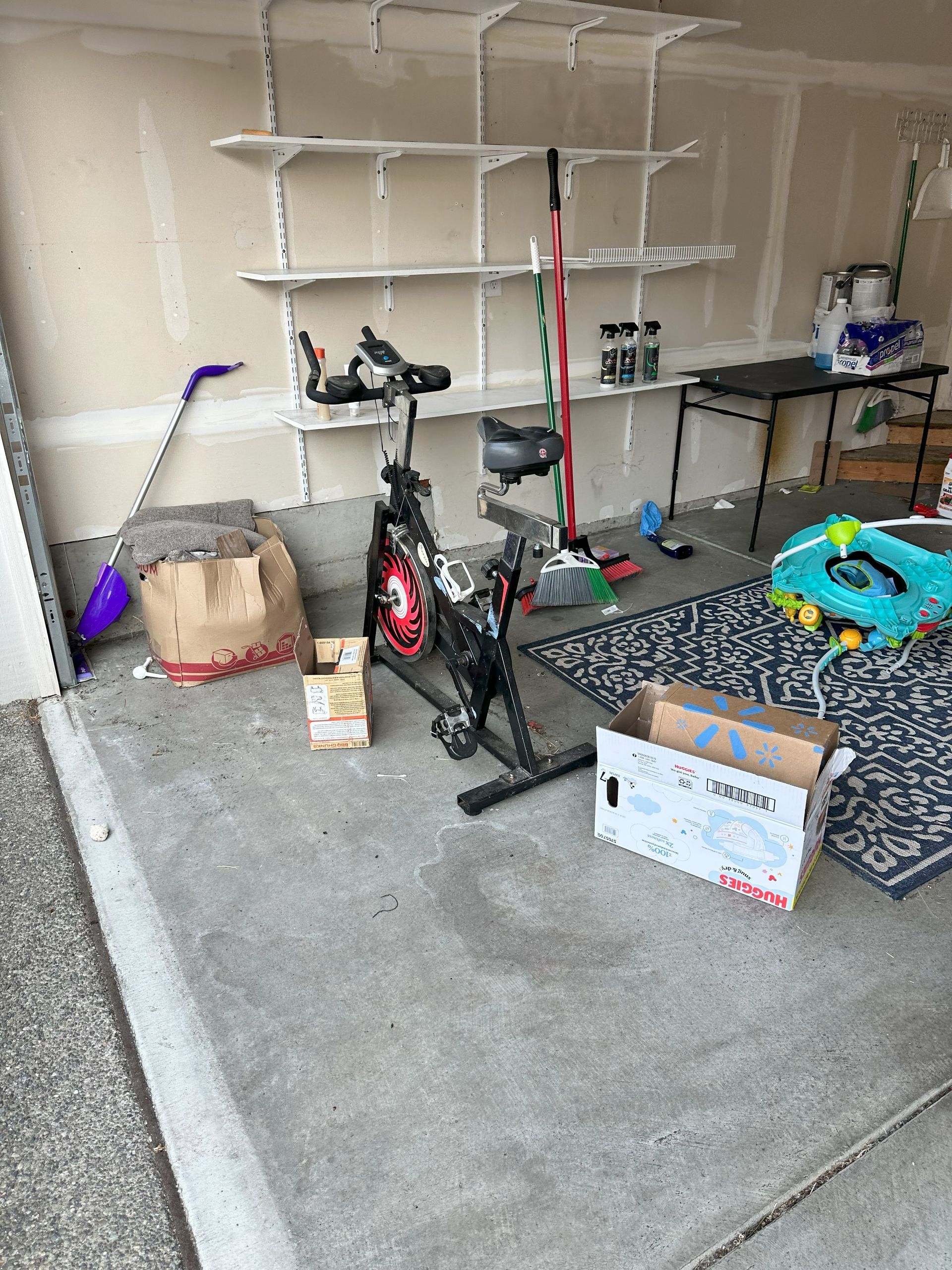 Garage with exercise bike, shelves, cardboard boxes, and a small table.