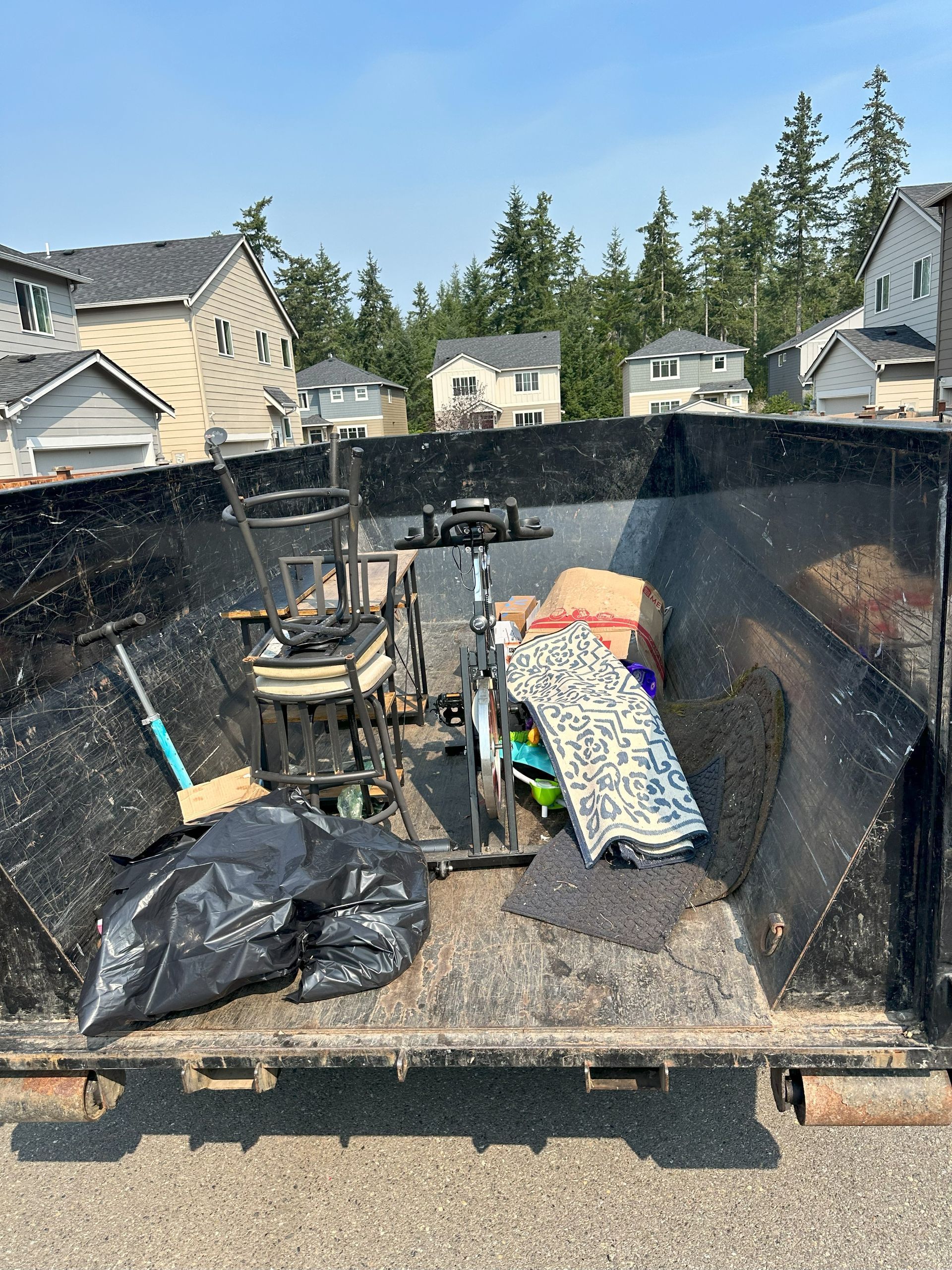 Dumpster filled with trash: black bags, rug, chair, and other discarded items. Houses and trees in the background.