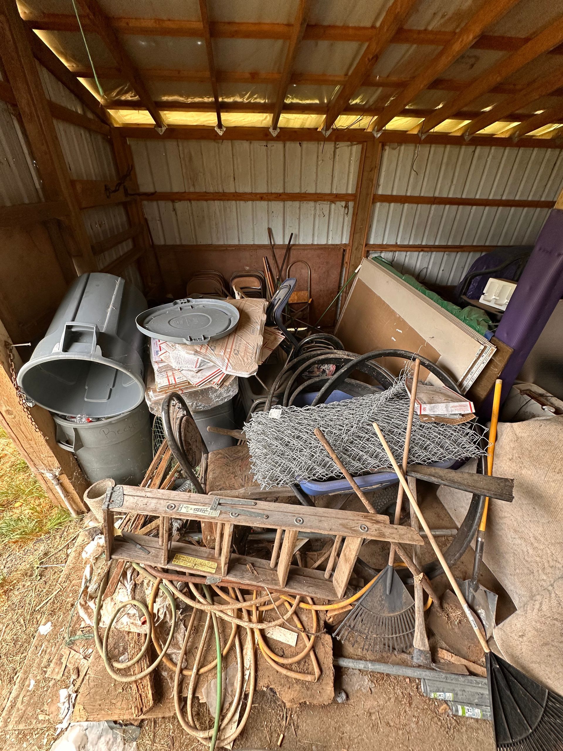 A cluttered shed interior, filled with items like a ladder, trash can, and rolled wire.