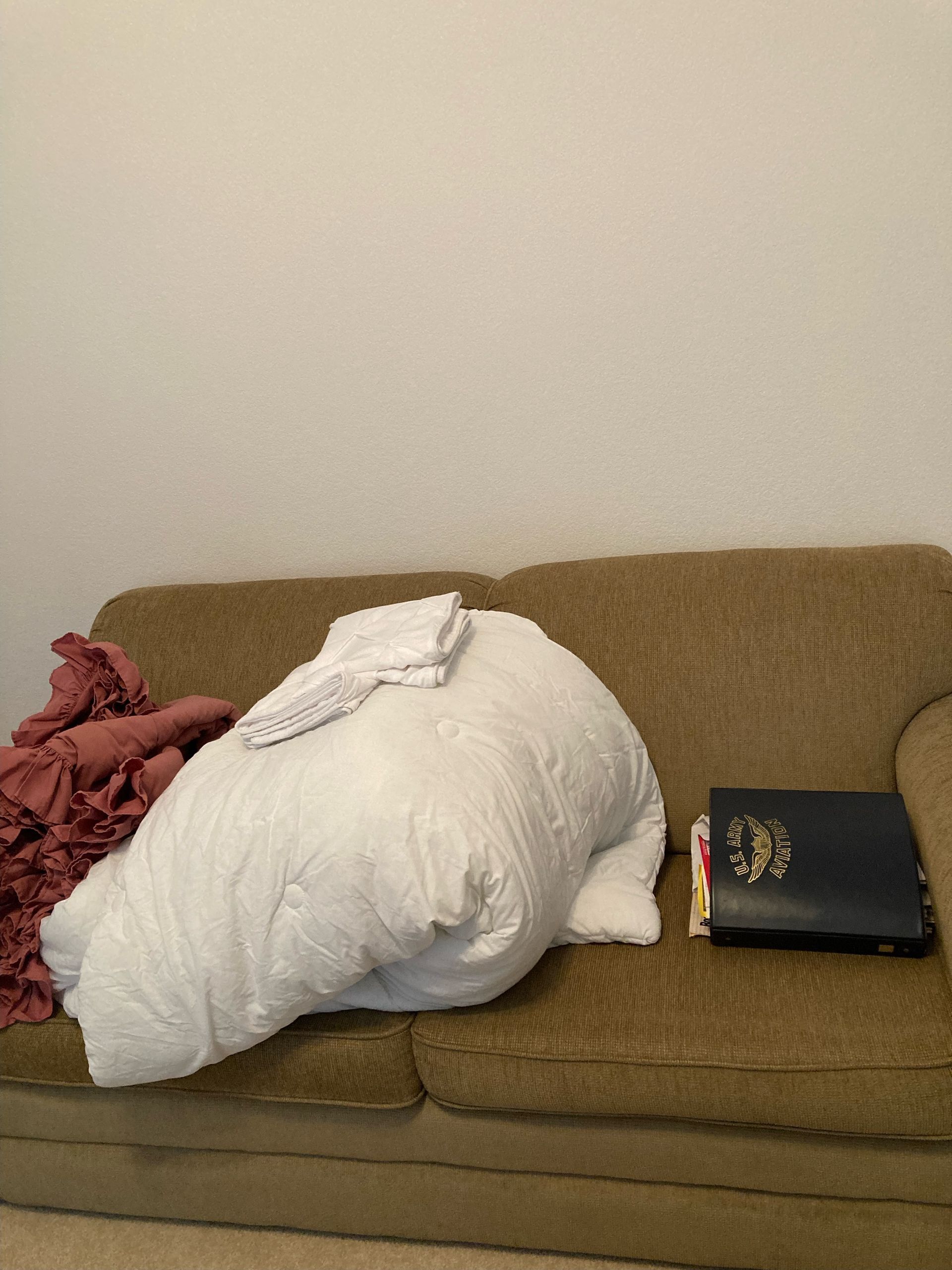A brown couch with a large white comforter, a pink blanket, and a black binder.