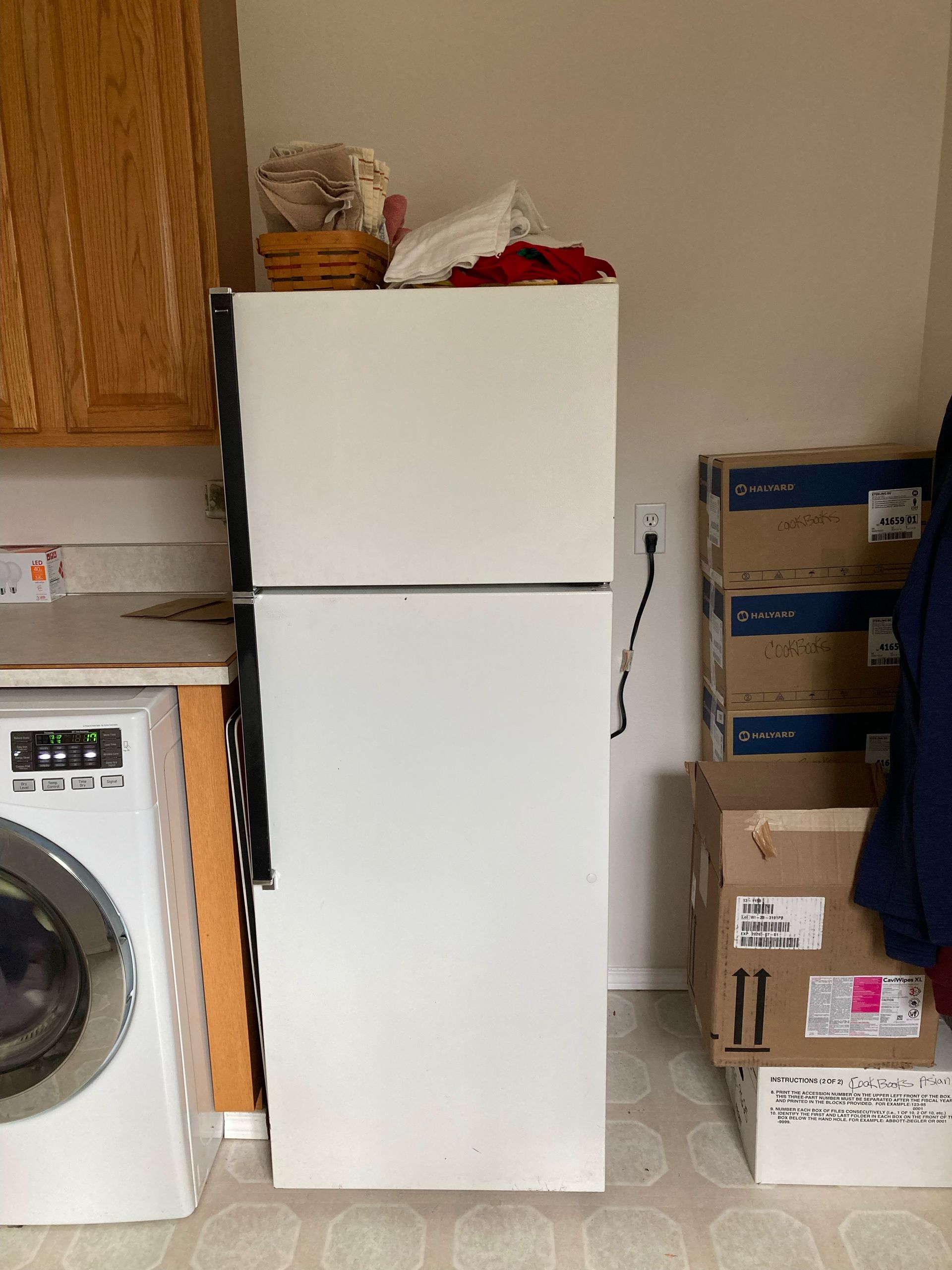 White refrigerator next to cabinets, washing machine, and stacked boxes in a room.
