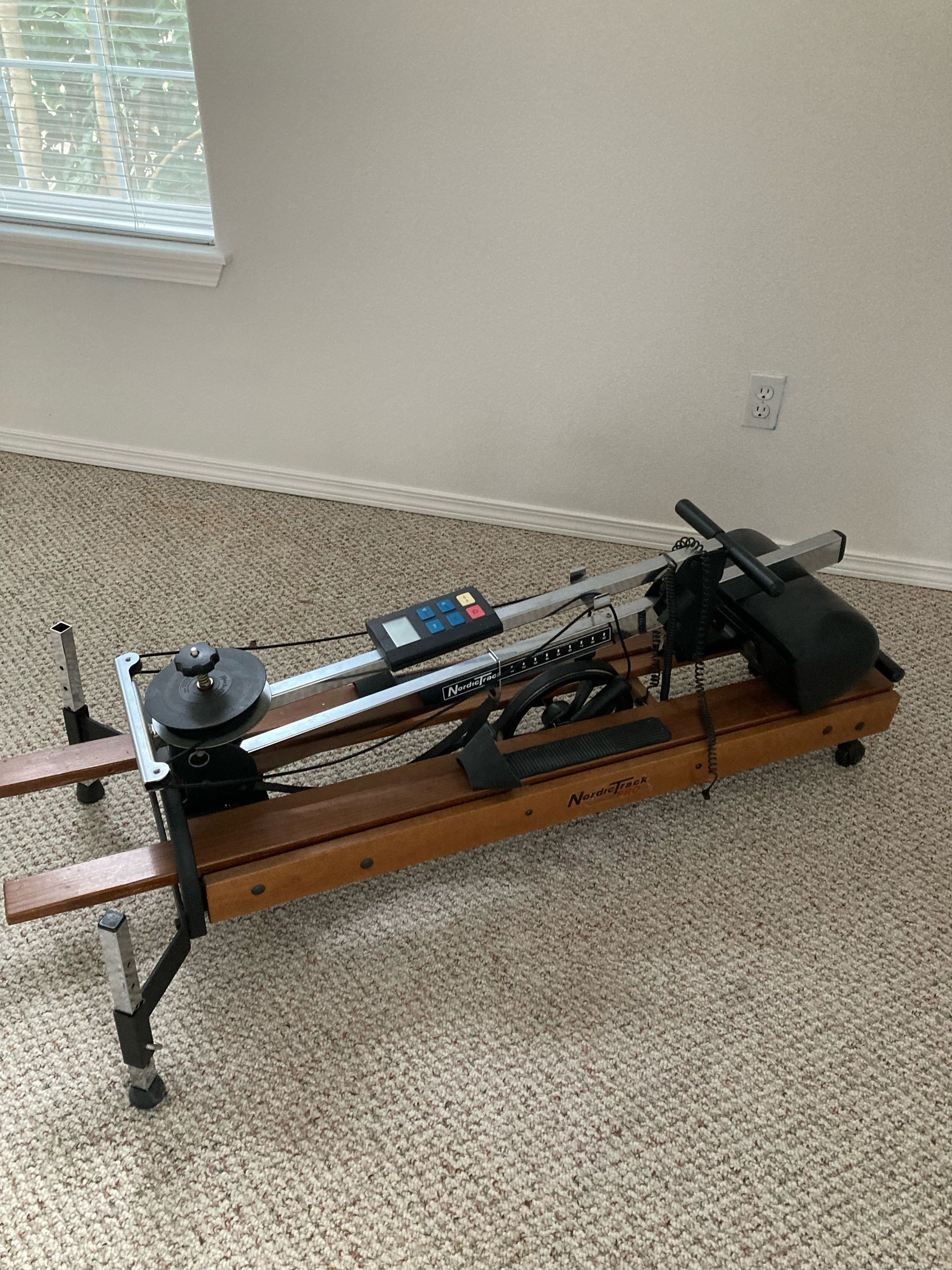 Rowing machine on a tan carpet, near a window. It has a digital display, a wooden frame, and black components.