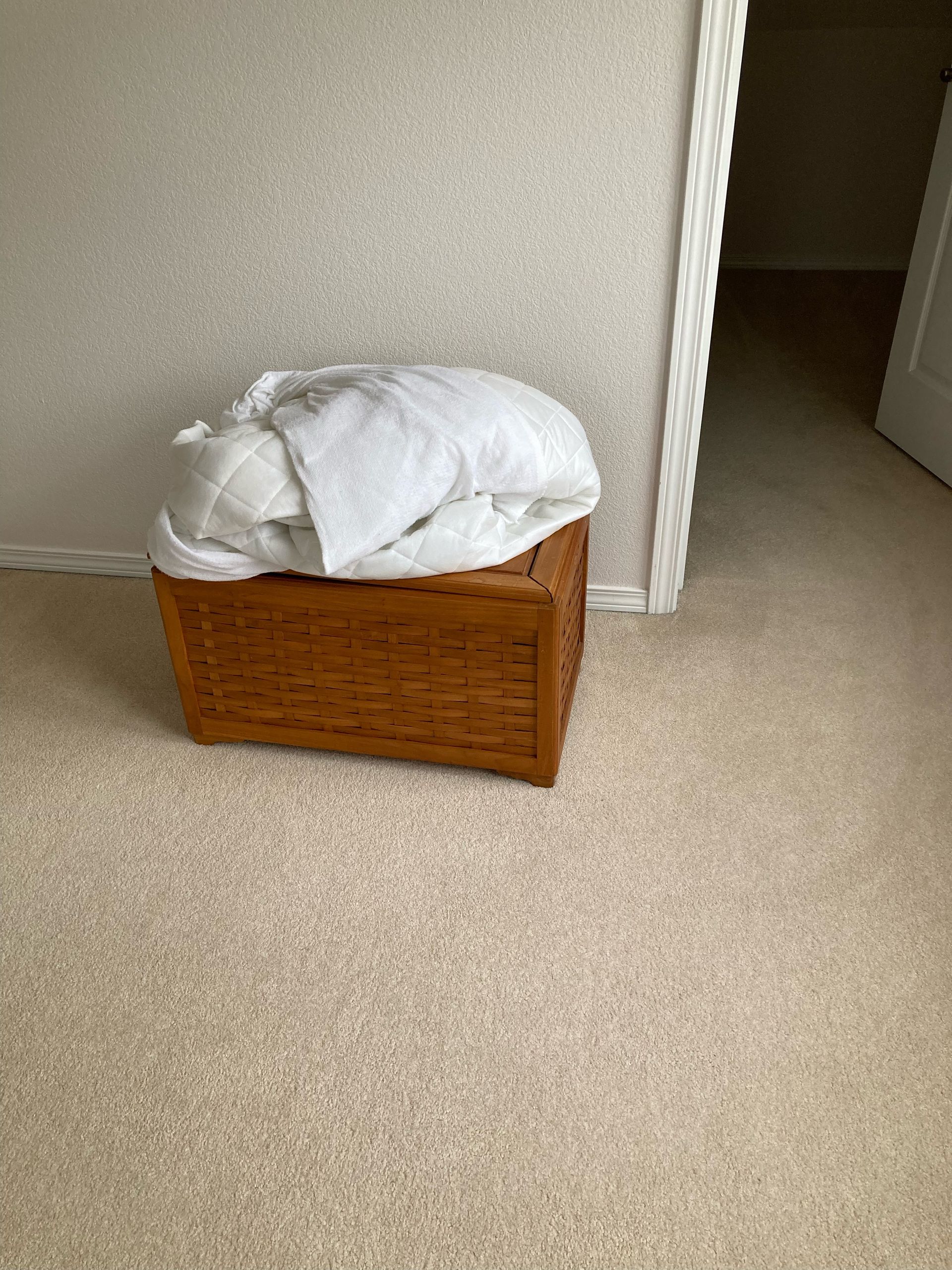 A woven wooden trunk with white bedding sits against a wall in a room with beige carpet.