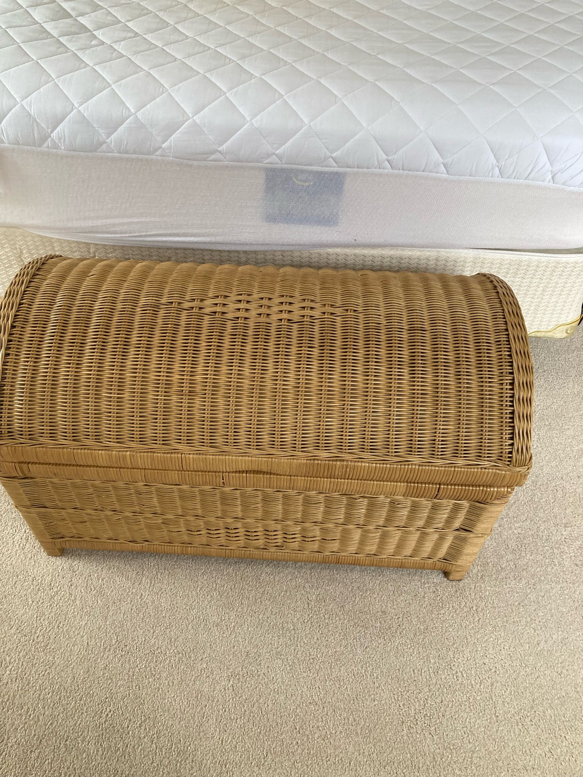 Wicker chest at the foot of a bed on a beige carpet.