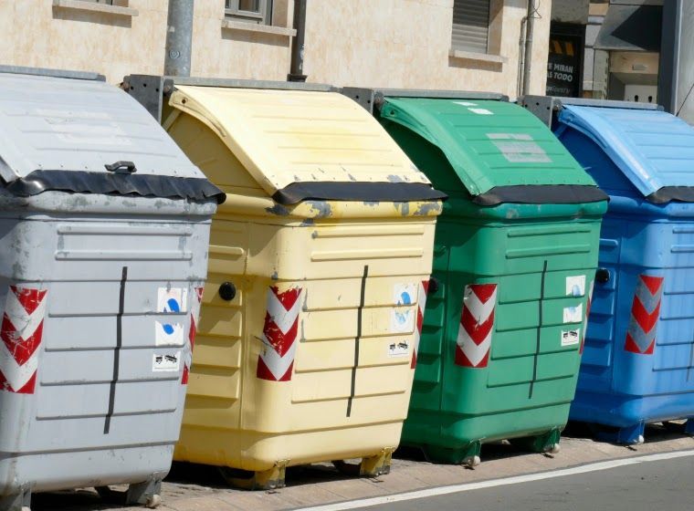 Four colorful recycling bins: gray, yellow, green, and blue, lined up on a street.