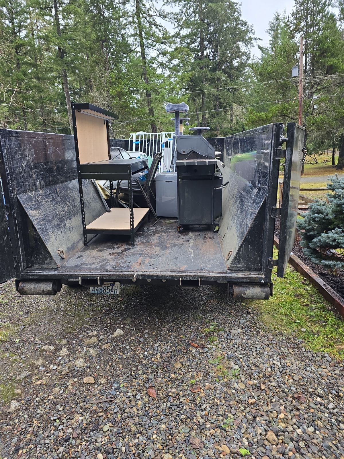 A black dumpster filled with discarded items: grill, shelving, and a refrigerator, outdoors.