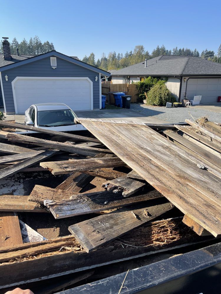 Pile of weathered wood boards in a truck bed, houses and car in the background on a sunny day.