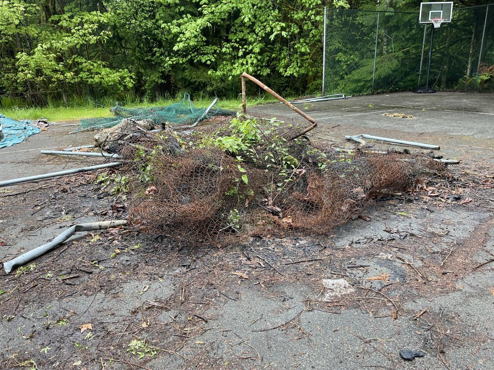 Debris pile on asphalt; includes metal fencing, vegetation, and a basketball hoop in the background.