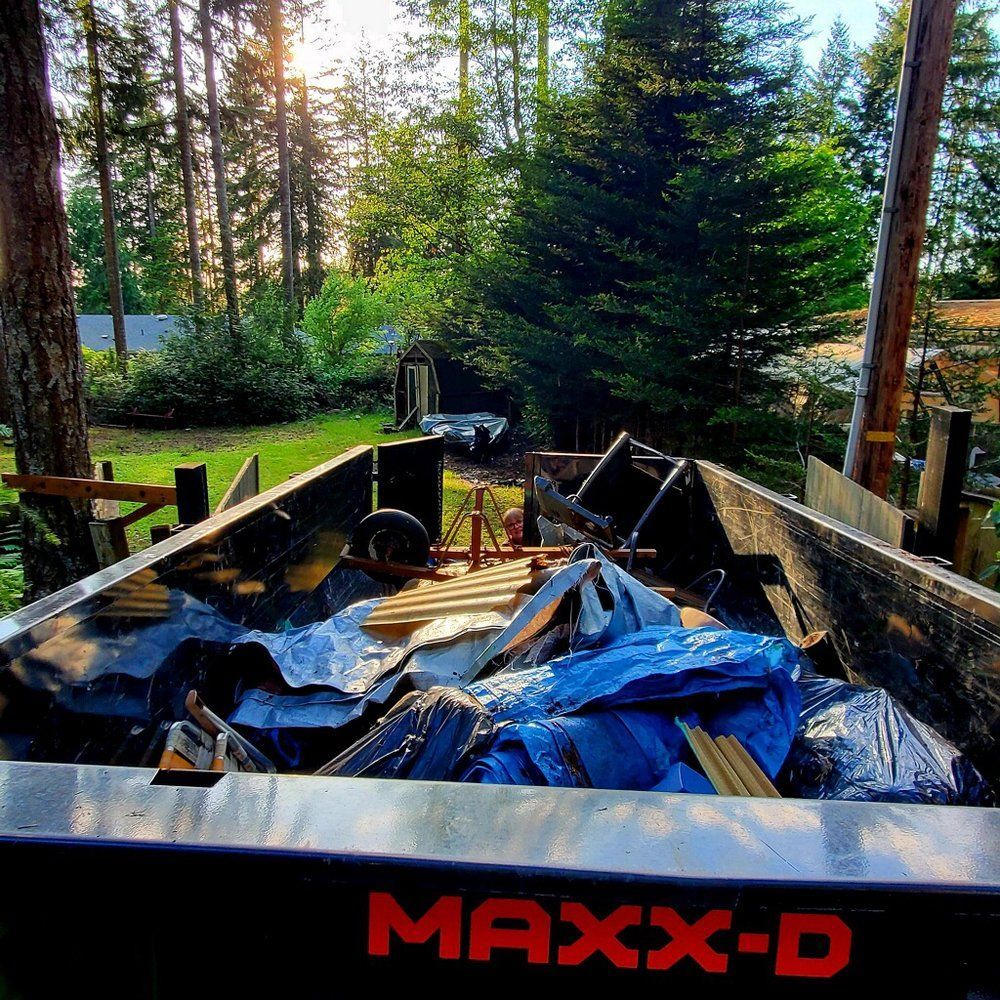 A loaded black MAX-D trailer in a wooded backyard, containing scrap wood, metal, and blue tarps.