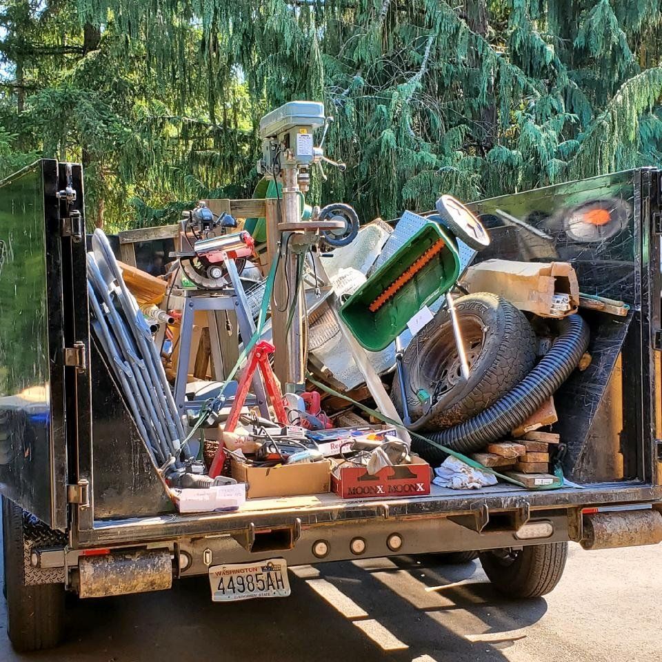 A pickup truck bed overflowing with tools, scrap wood, a tire, and yard waste, under sunny trees.
