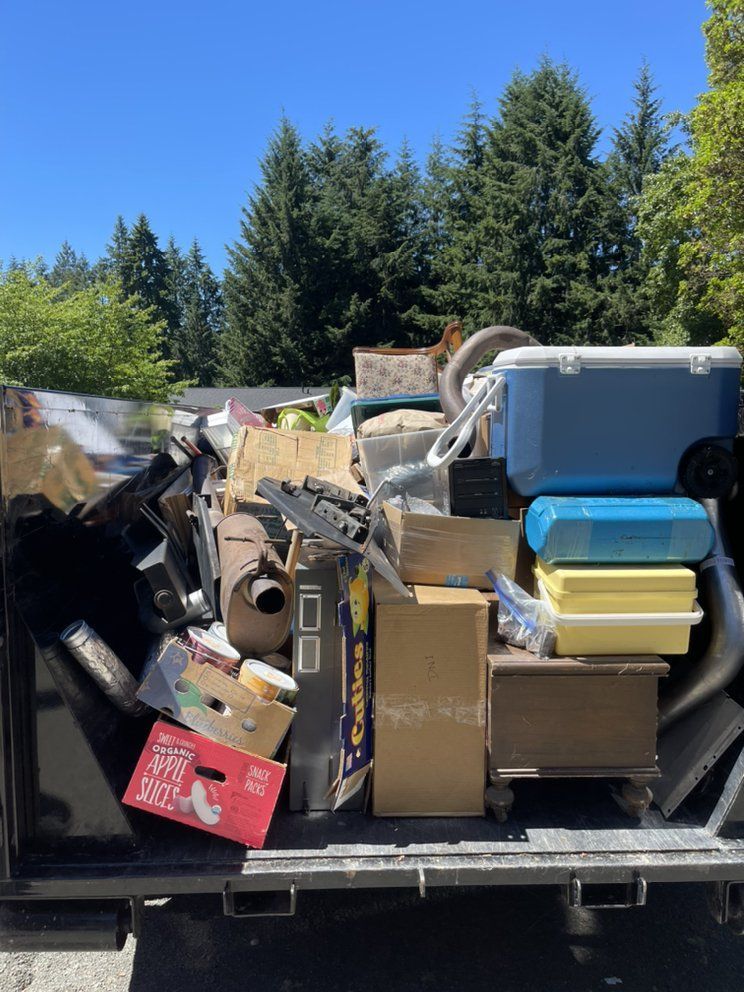 Dumpster filled with trash under a blue sky. Items include boxes, cooler, and various debris.